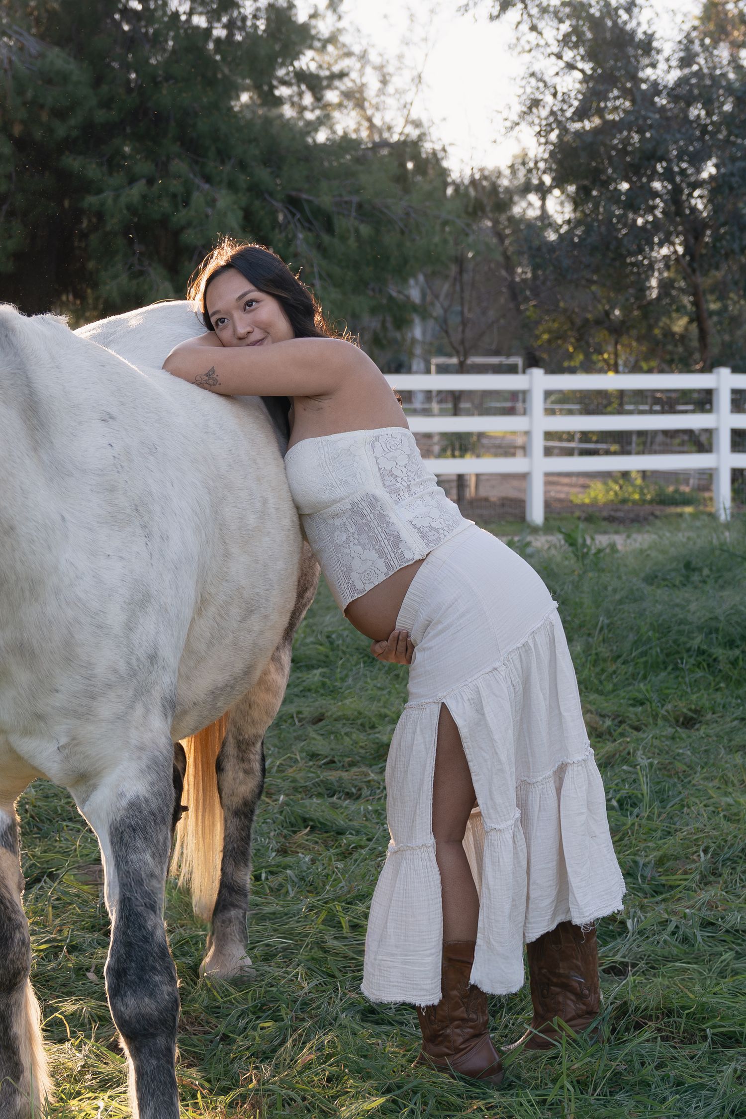 Young woman in white dress affectionately hugs white horse at sunny farm setting.