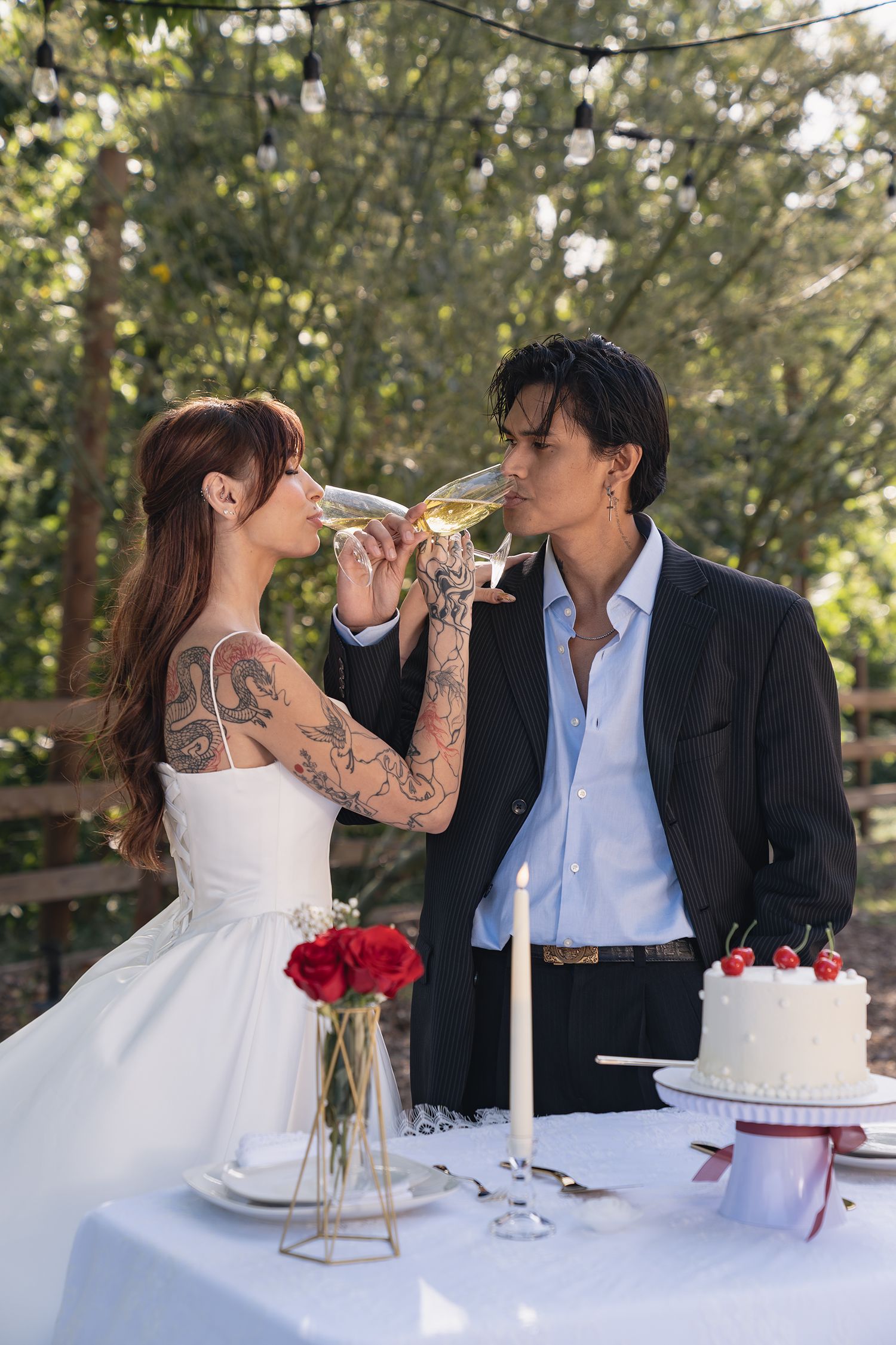 A tattooed bride and groom toast with champagne glasses at an outdoor wedding reception table.