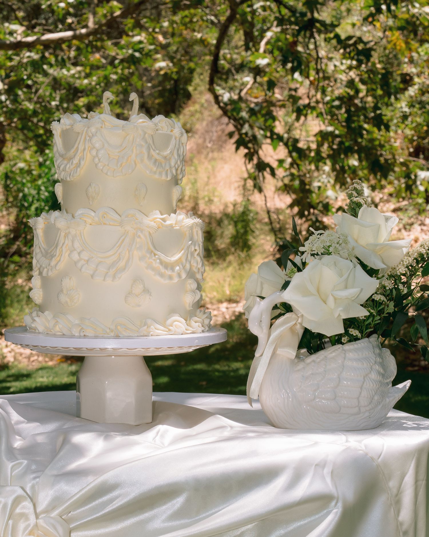 A two-tier white wedding cake with piped details sits beside a swan vase with white roses.
