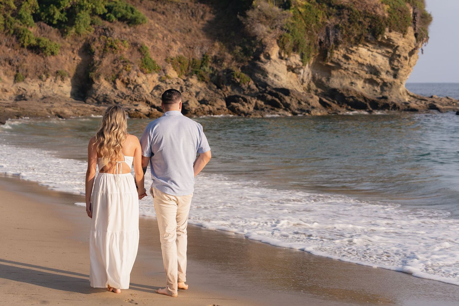 A couple walks hand-in-hand along a scenic beach with rocky cliffs at sunset.
