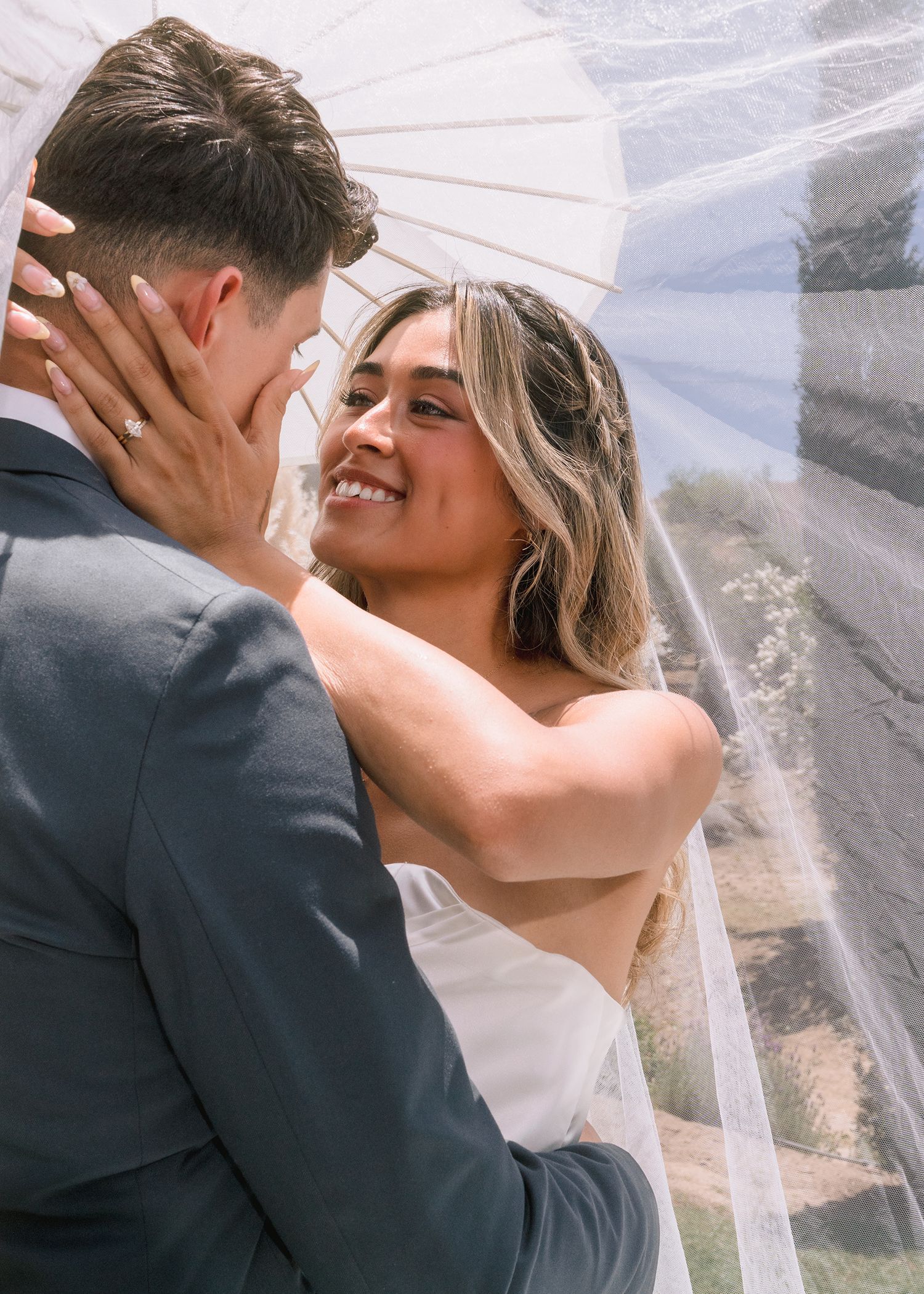 Smiling bride in white gown embraces groom under a veil on a sunny wedding day.