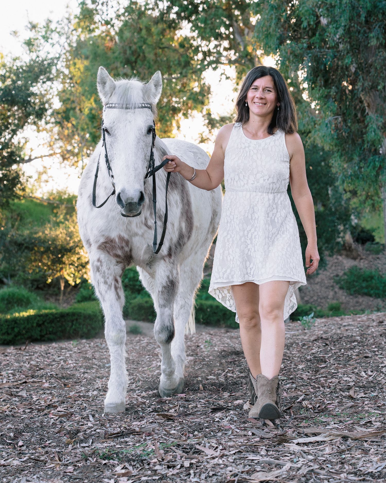 Woman in white lace dress walking with a white Appaloosa horse in a sunlit outdoor setting.