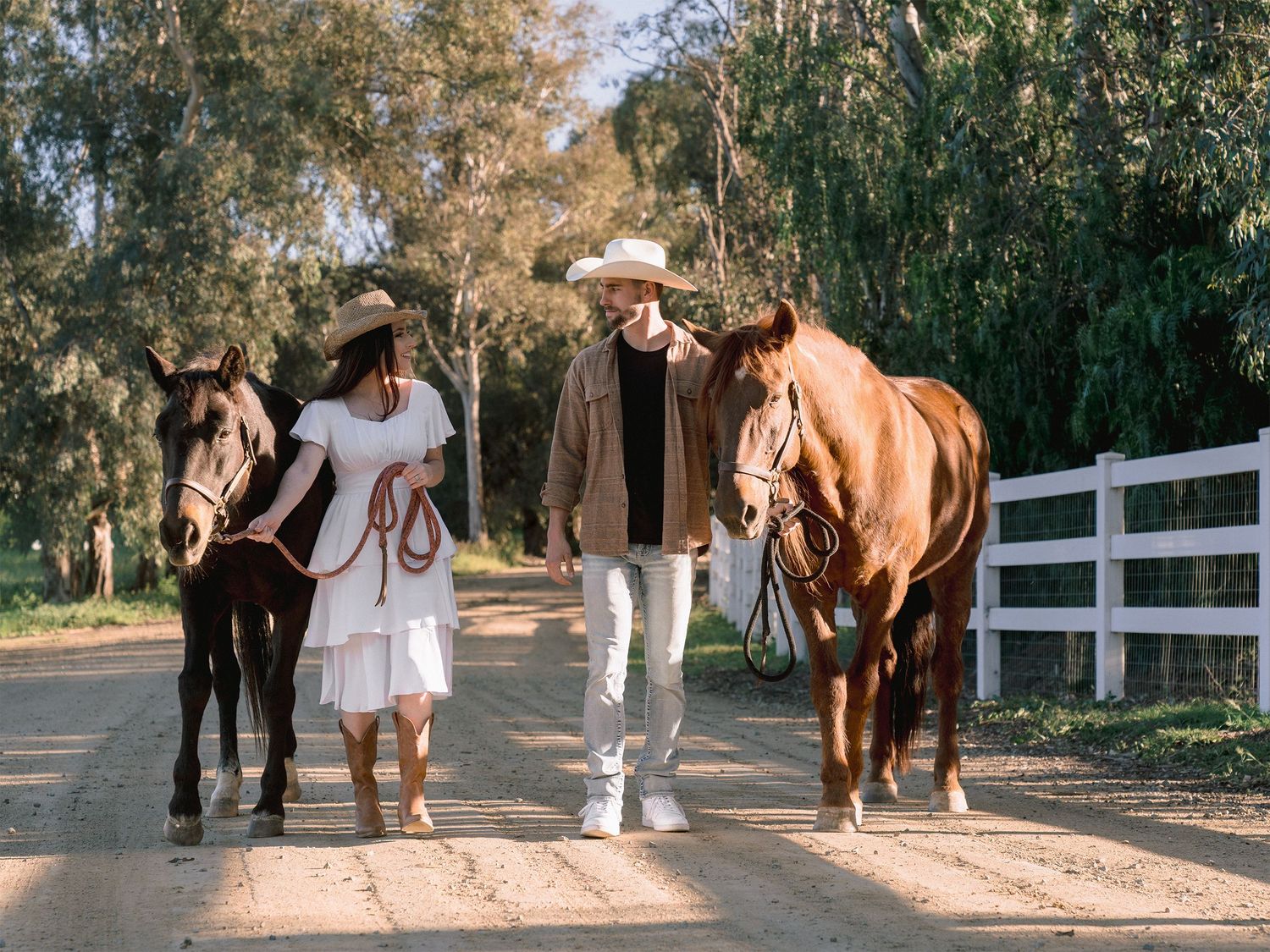 Couple walking two horses on a dirt path with white fence and trees in a sunny outdoor setting.