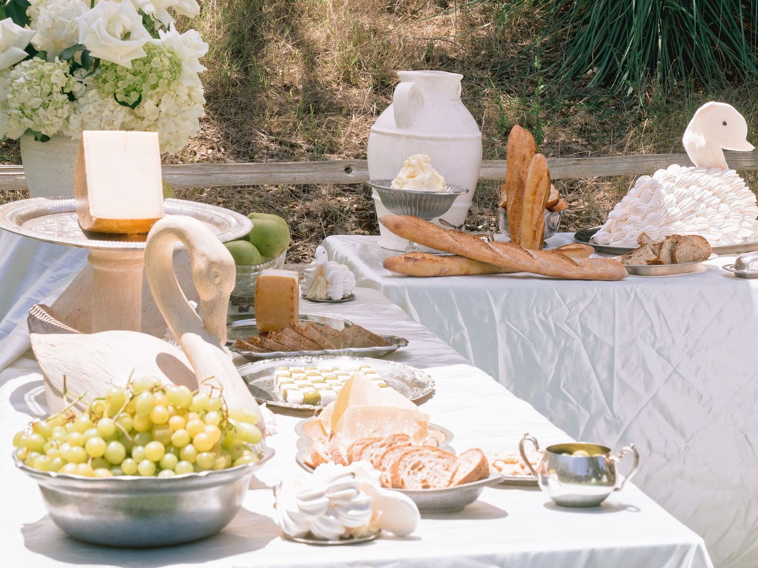 An elegant outdoor wedding table displays grapes, bread, cheeses, and white floral arrangements.