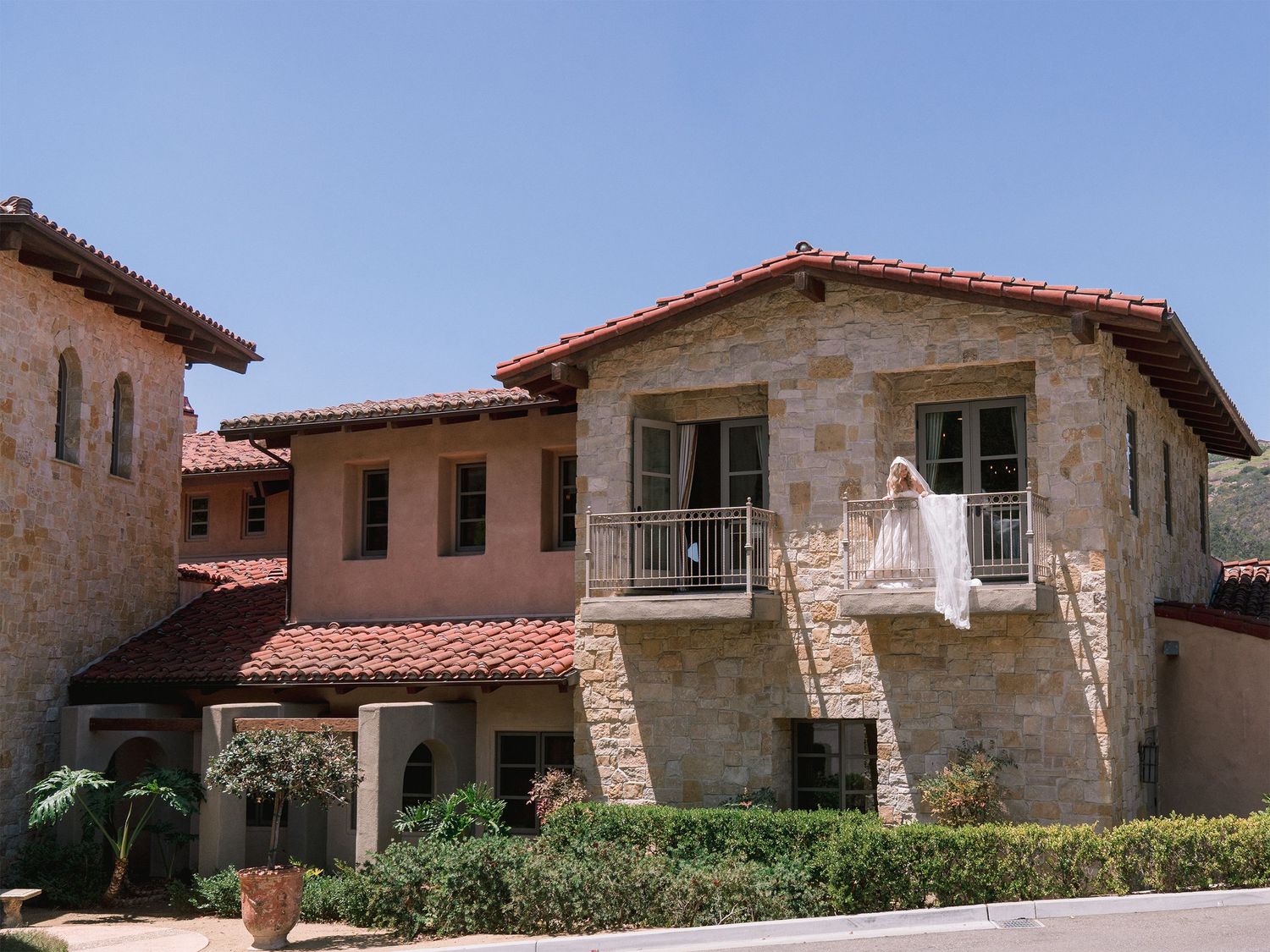 A wedding dress hangs from a balcony of a Spanish-style stone villa with red tile roofs.