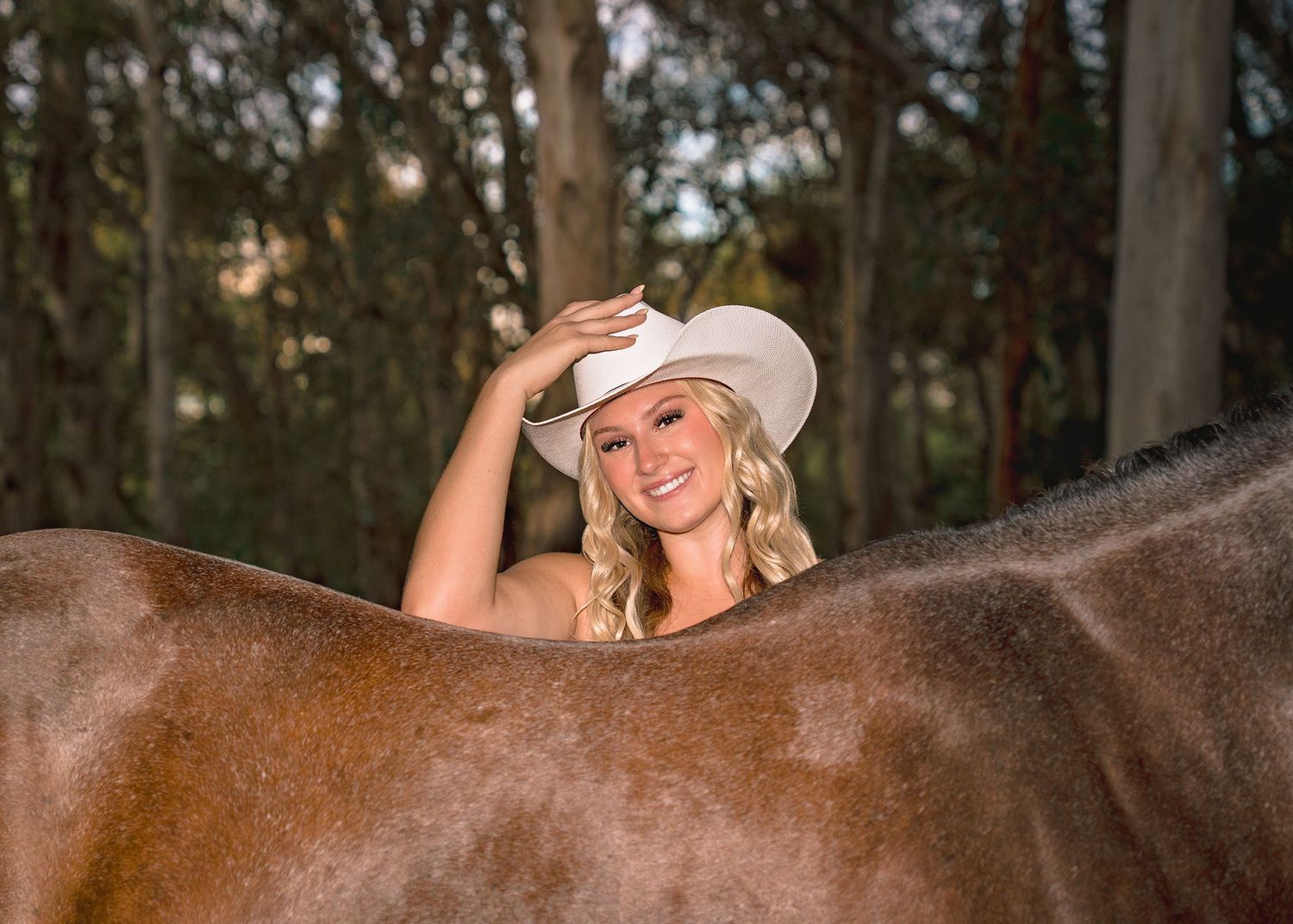 Smiling blonde woman in white cowboy hat leaning over a brown horse in a forest setting.