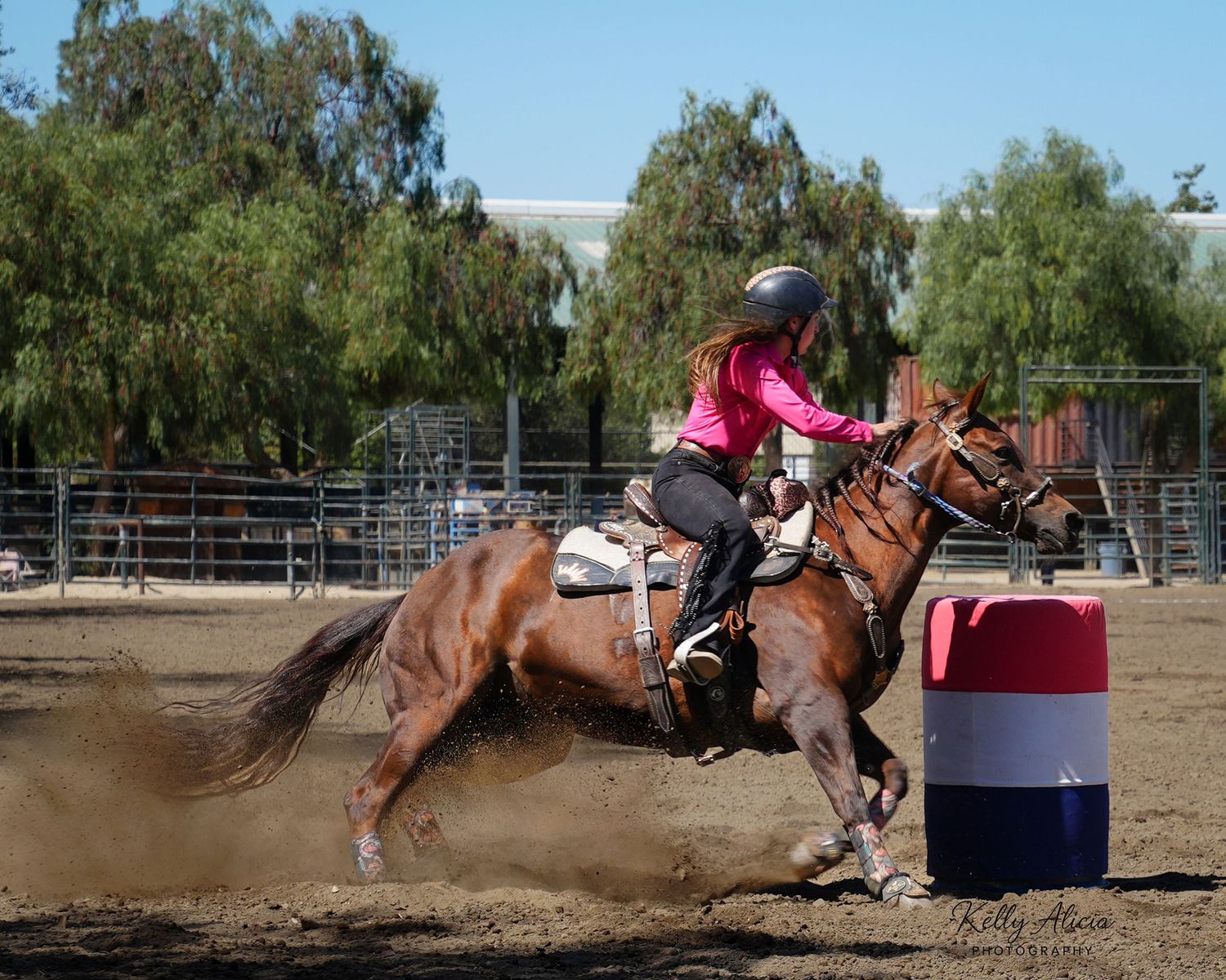 Female rider in pink shirt racing a chestnut horse around a barrel in a sandy rodeo arena.