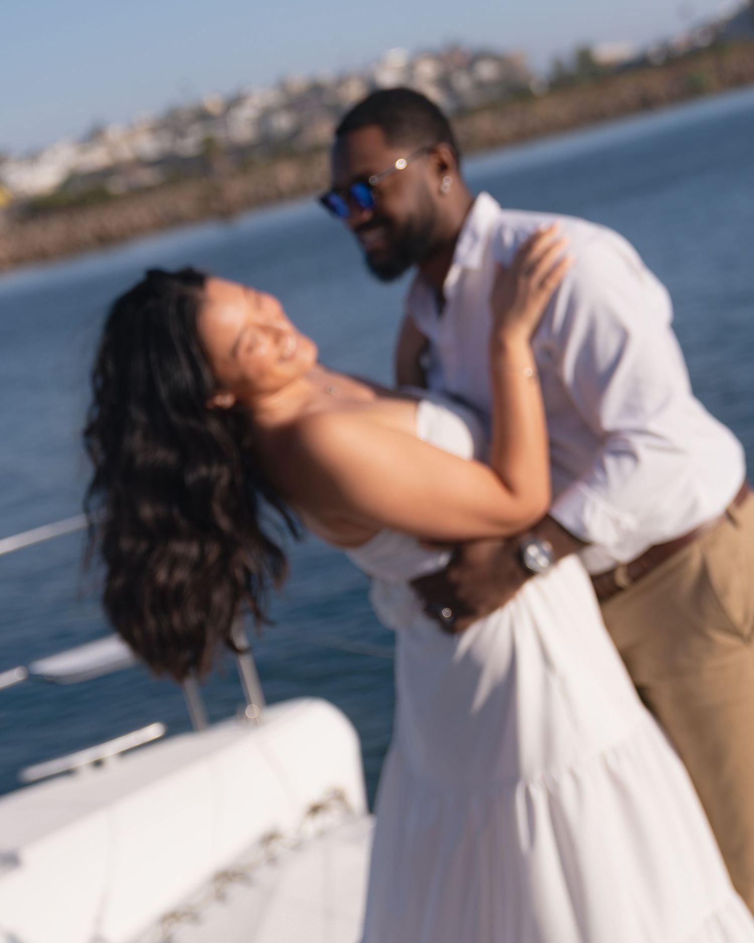 Couple in white outfits share a joyful dip pose on a boat deck, with the sparkling blue sea and a sunlit coastline behind them.