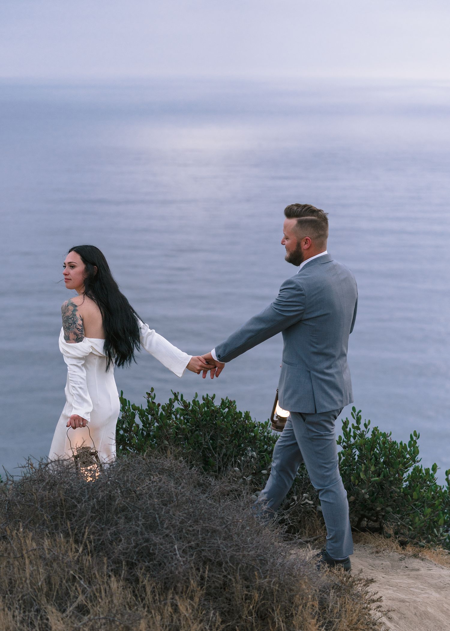 Couple holding hands on a coastal cliff, woman in white dress with tattoos, man in gray suit, ocean backdrop.