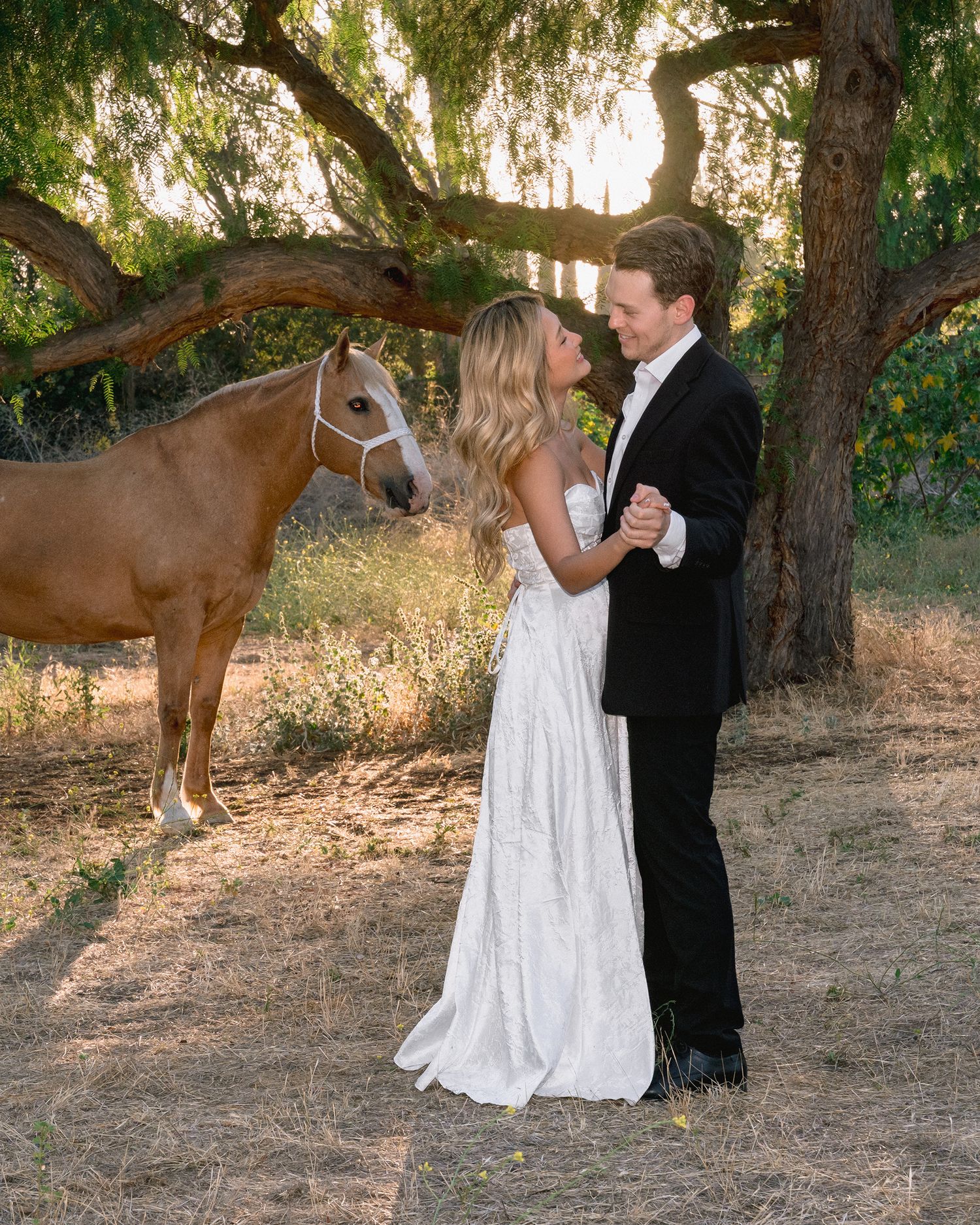 A bride in a flowing white gown and a groom in a black suit share an embrace beneath a large tree beside a palomino horse.