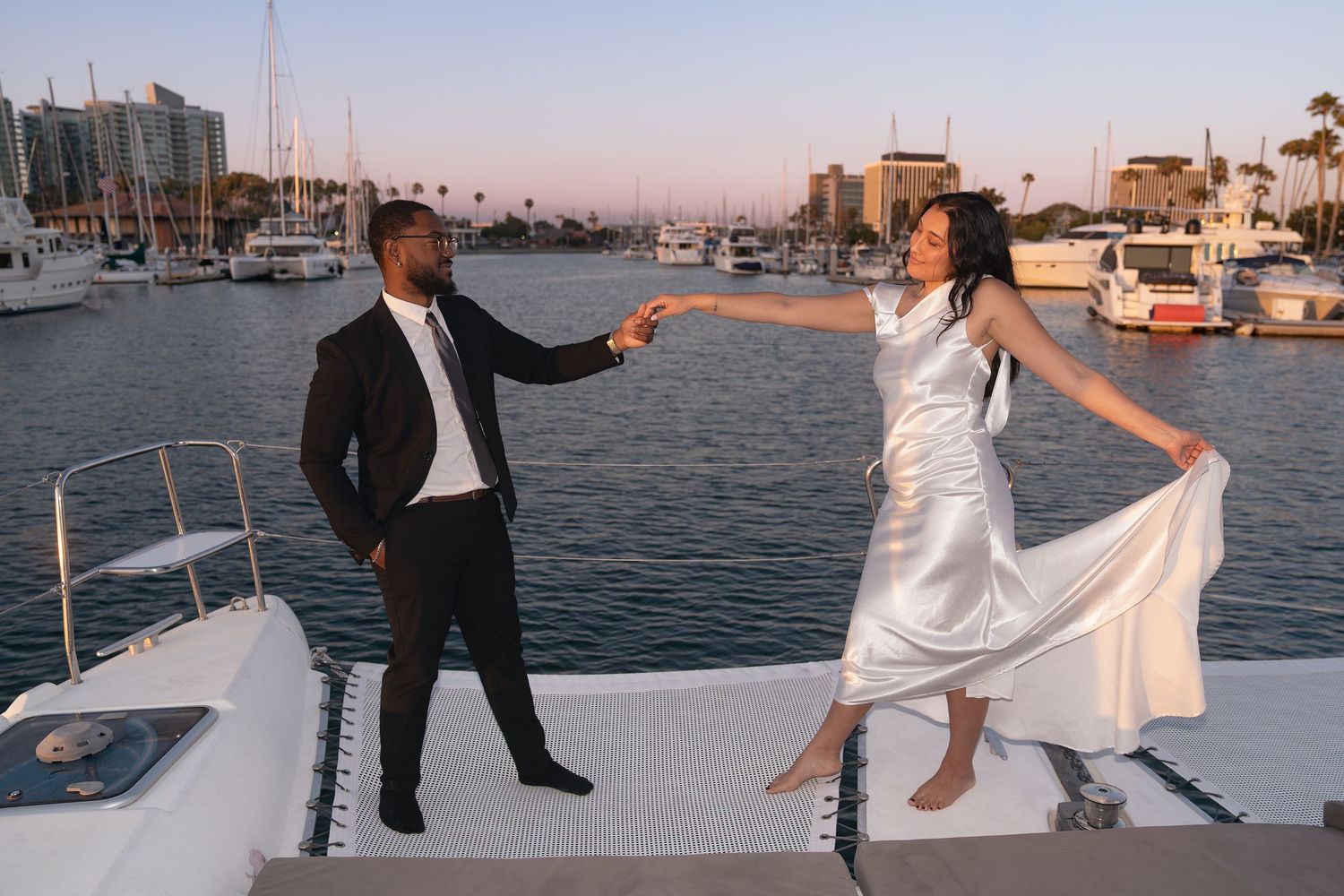 Couple in formal attire dancing hand in hand on a sailboat deck at sunset with a marina and boats in the background.