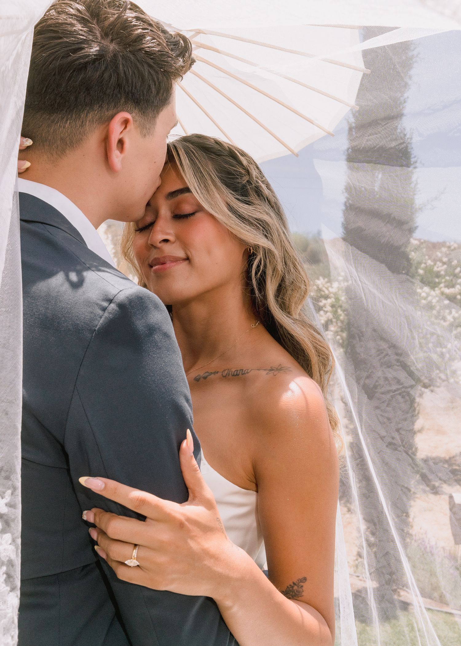 Bride in a white dress and groom in a gray suit embrace tenderly under a sheer veil and parasol, his lips resting on her forehead.