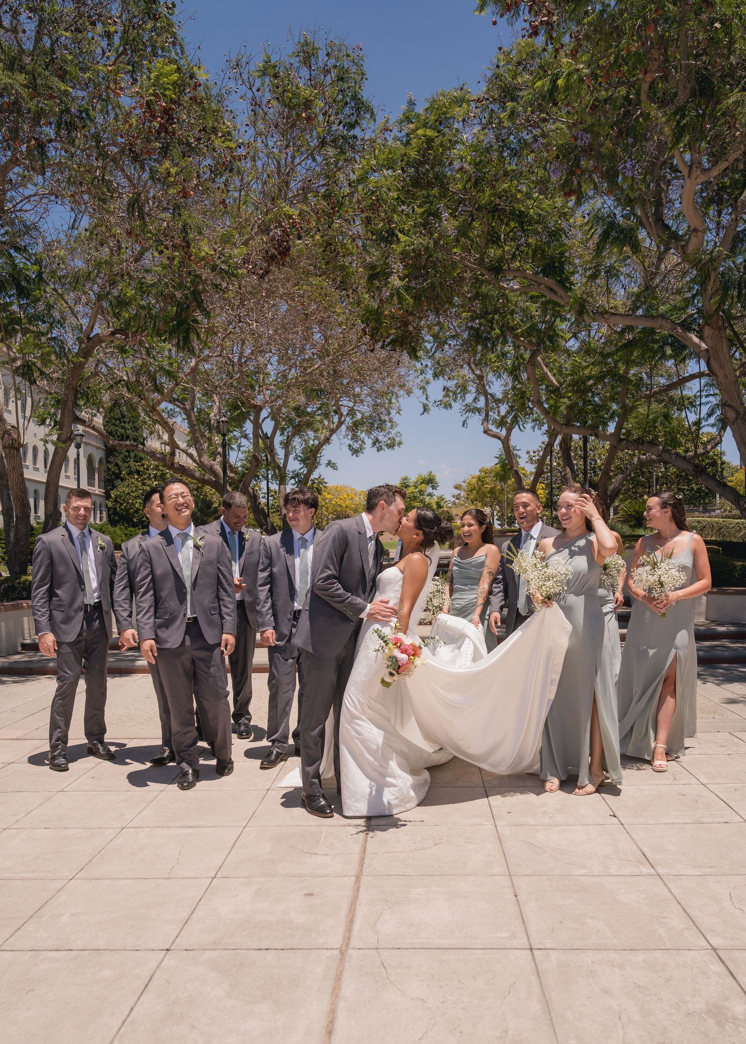 Wedding party gathered outdoors with bride and groom kissing, groomsmen in gray suits and bridesmaids in sage dresses.
