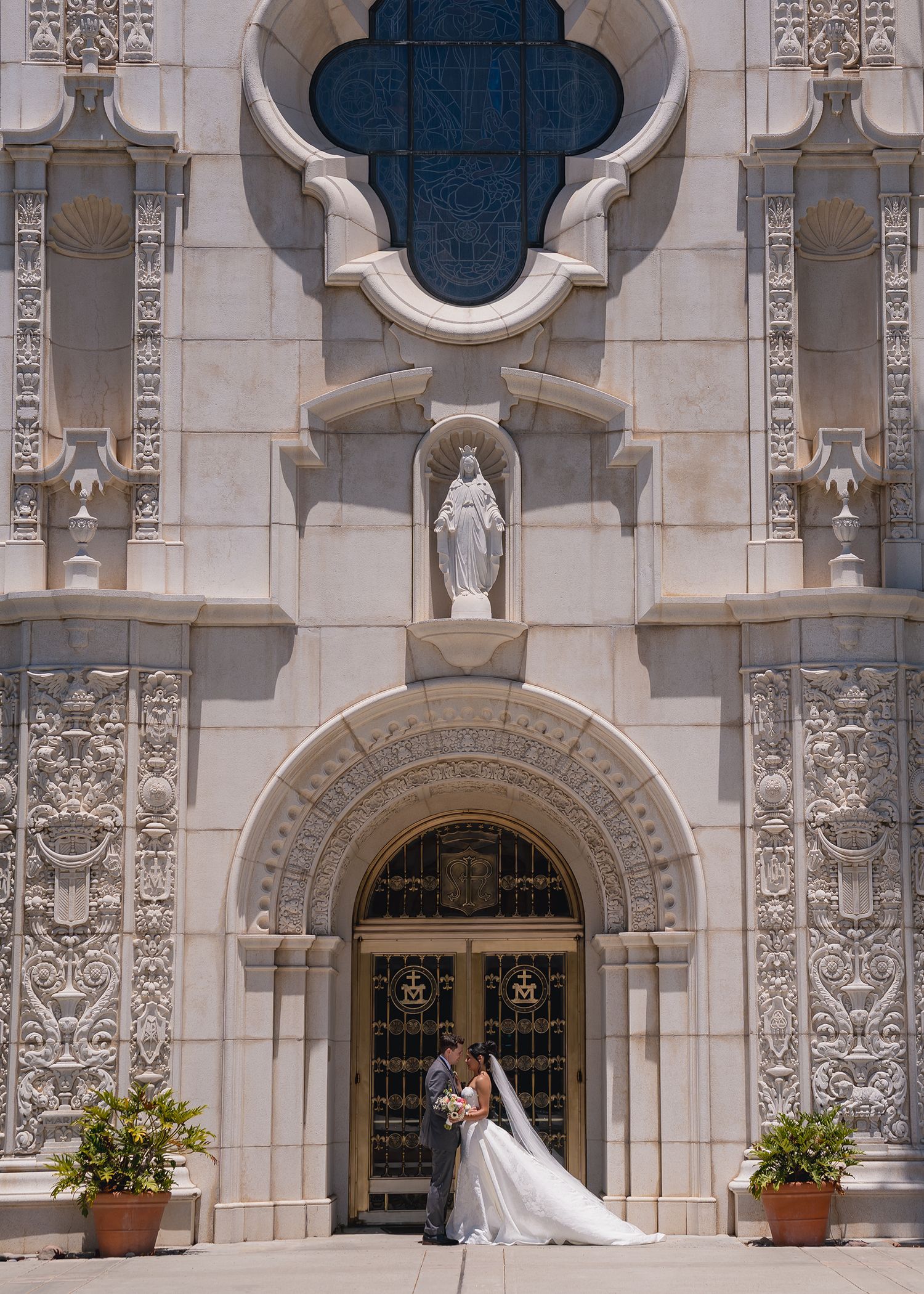 Bride and groom kissing at the ornate entrance of an elegant stone cathedral on their wedding day.