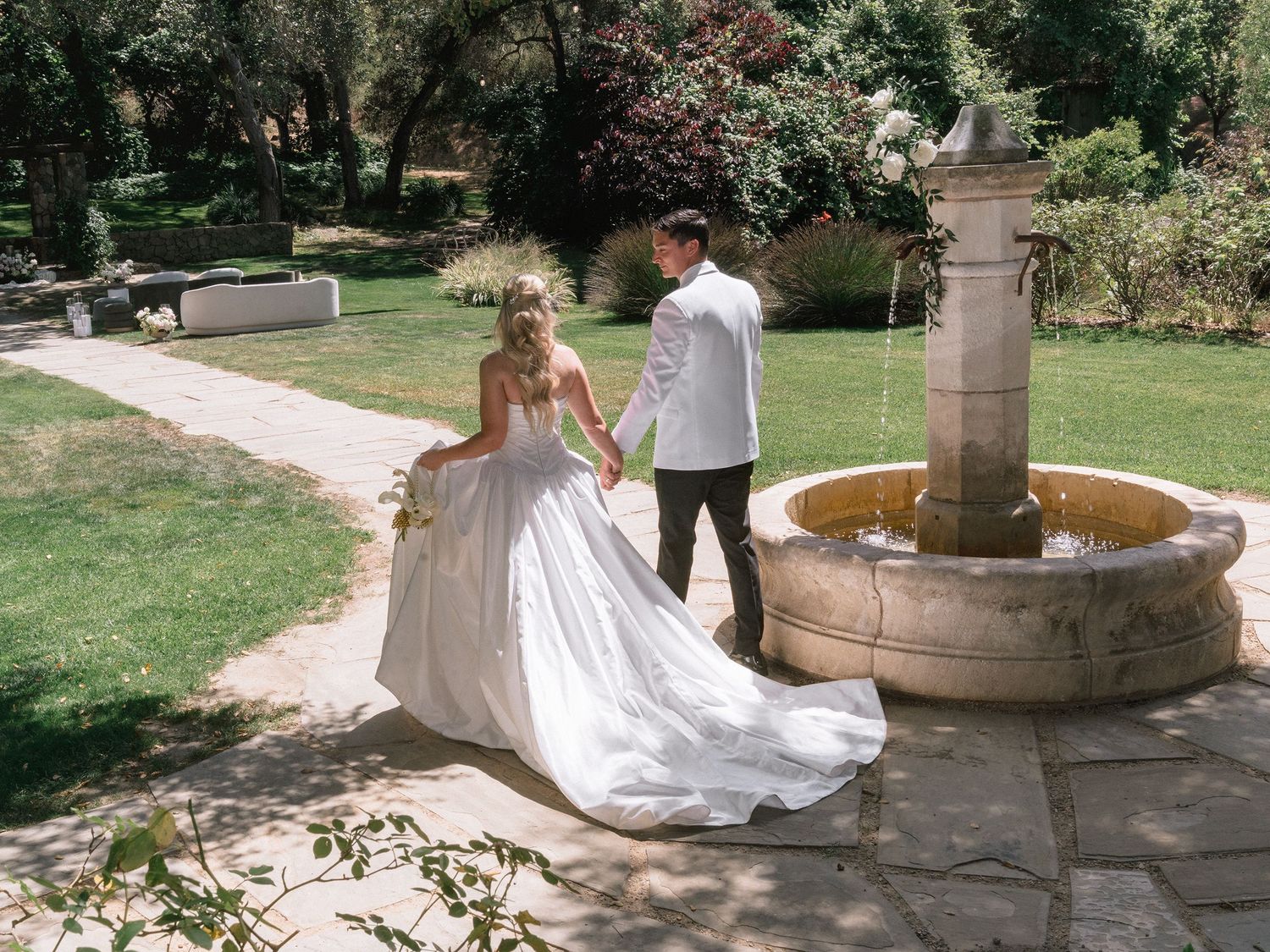 Bride in a flowing white gown and groom in a white shirt walk hand in hand past a stone fountain in a sunlit garden.