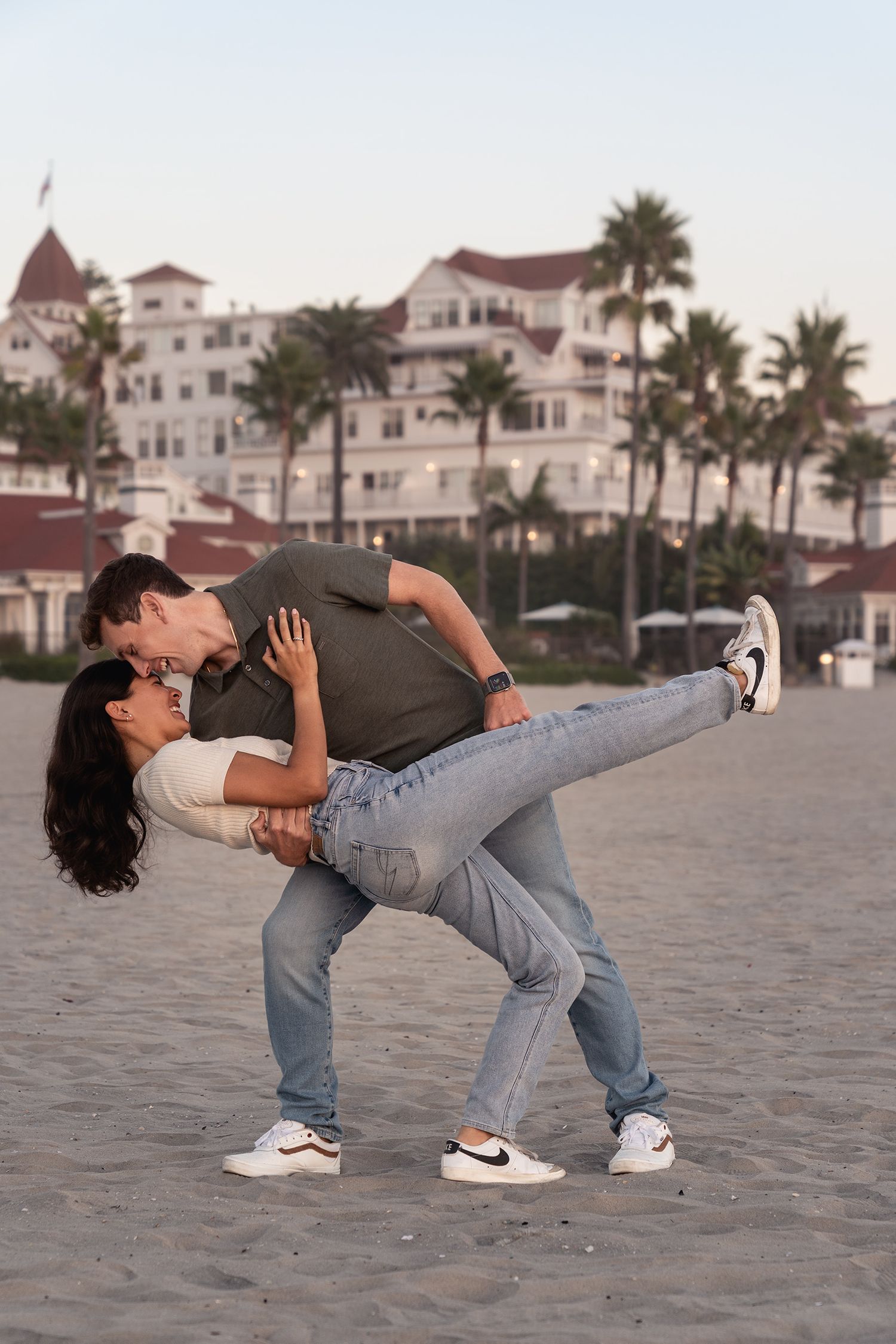 A couple shares a romantic dip kiss on a sandy beach with a grand hotel in the background.