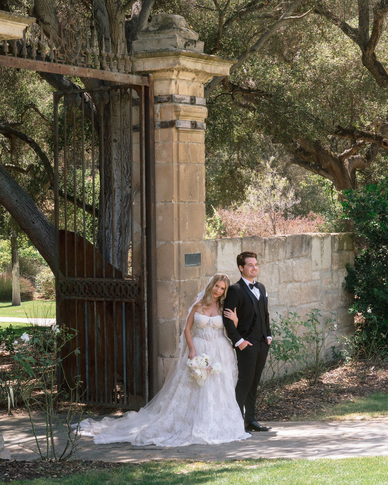 A bride in a white gown and groom in a tuxedo pose by a stone gate surrounded by oak trees.