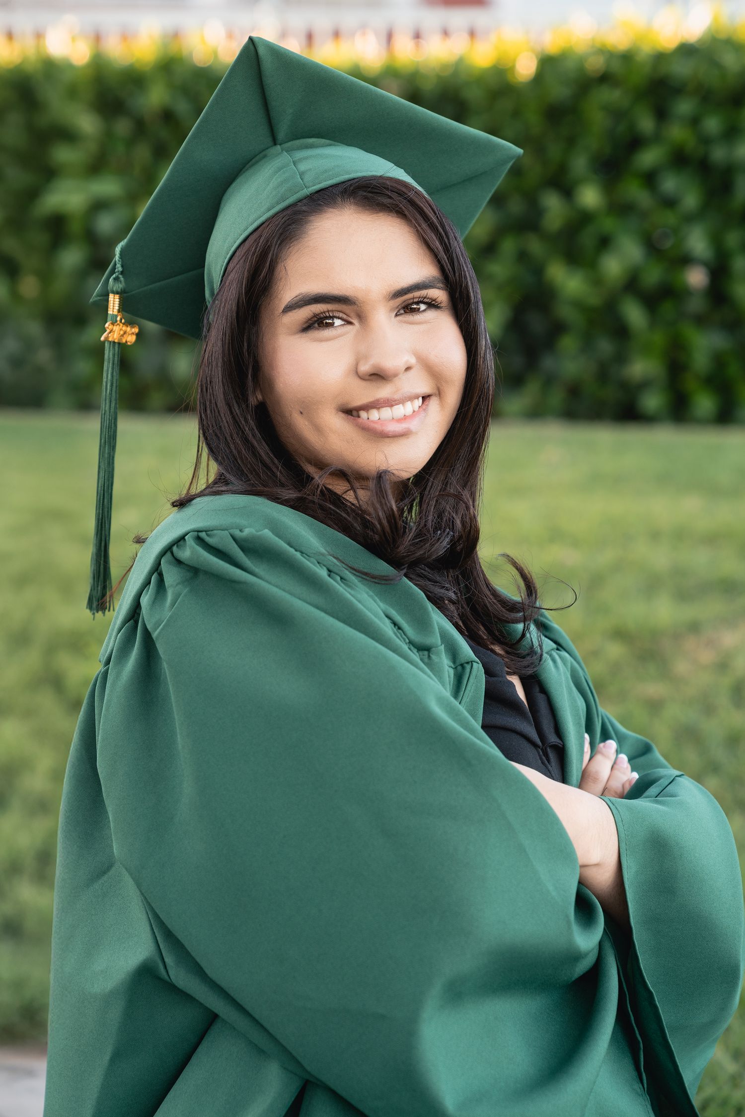 Smiling young woman in green graduation cap and gown poses with arms crossed outdoors.