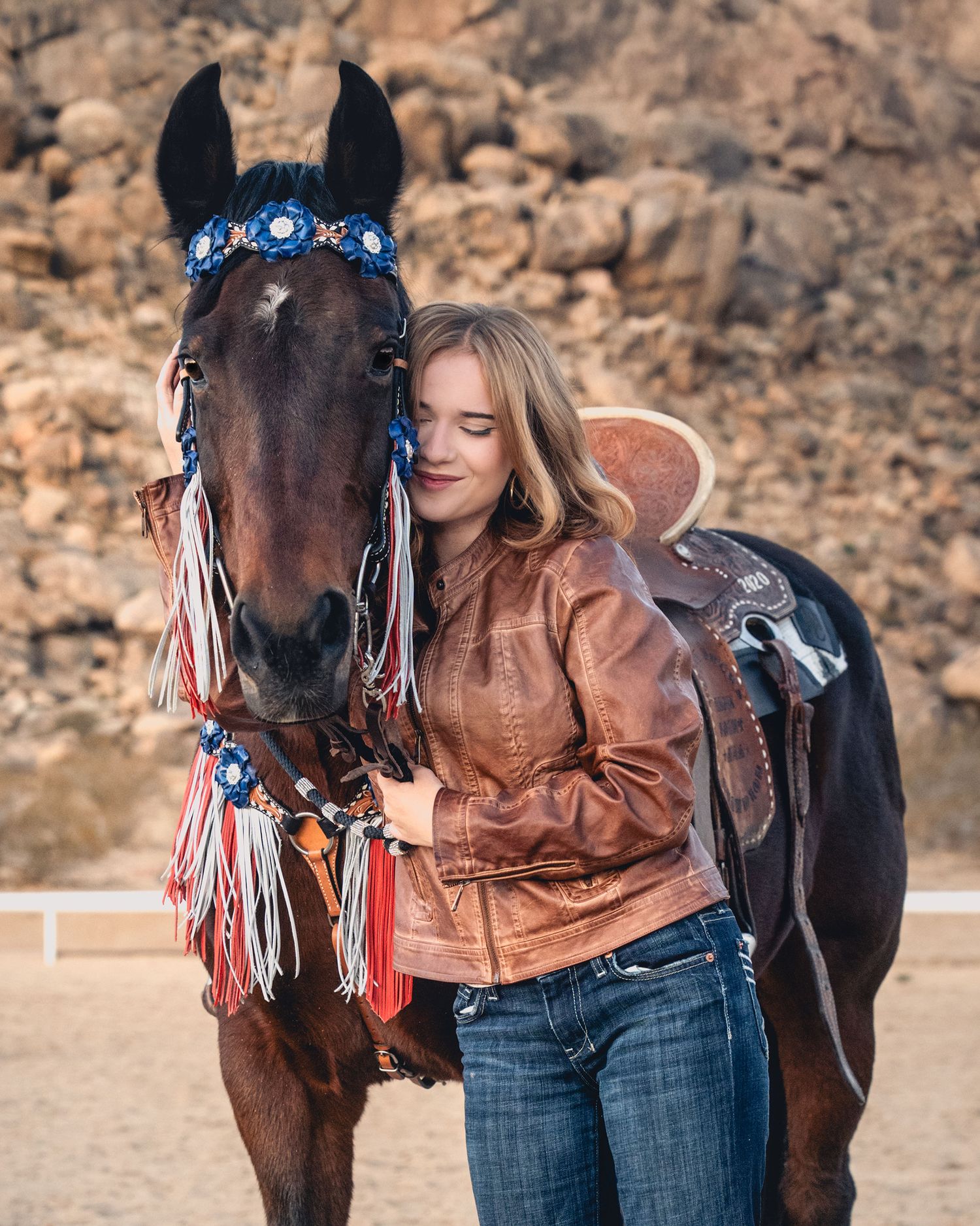 A blonde woman in a brown leather jacket affectionately poses with a decorated brown horse outdoors.