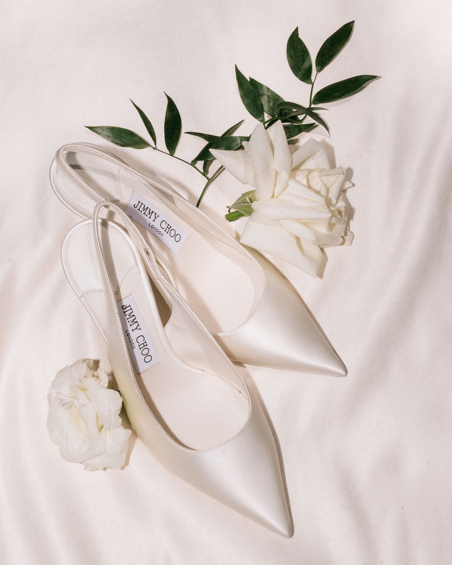 Jimmy Choo satin slingback bridal heels displayed with white flowers on a soft fabric background.