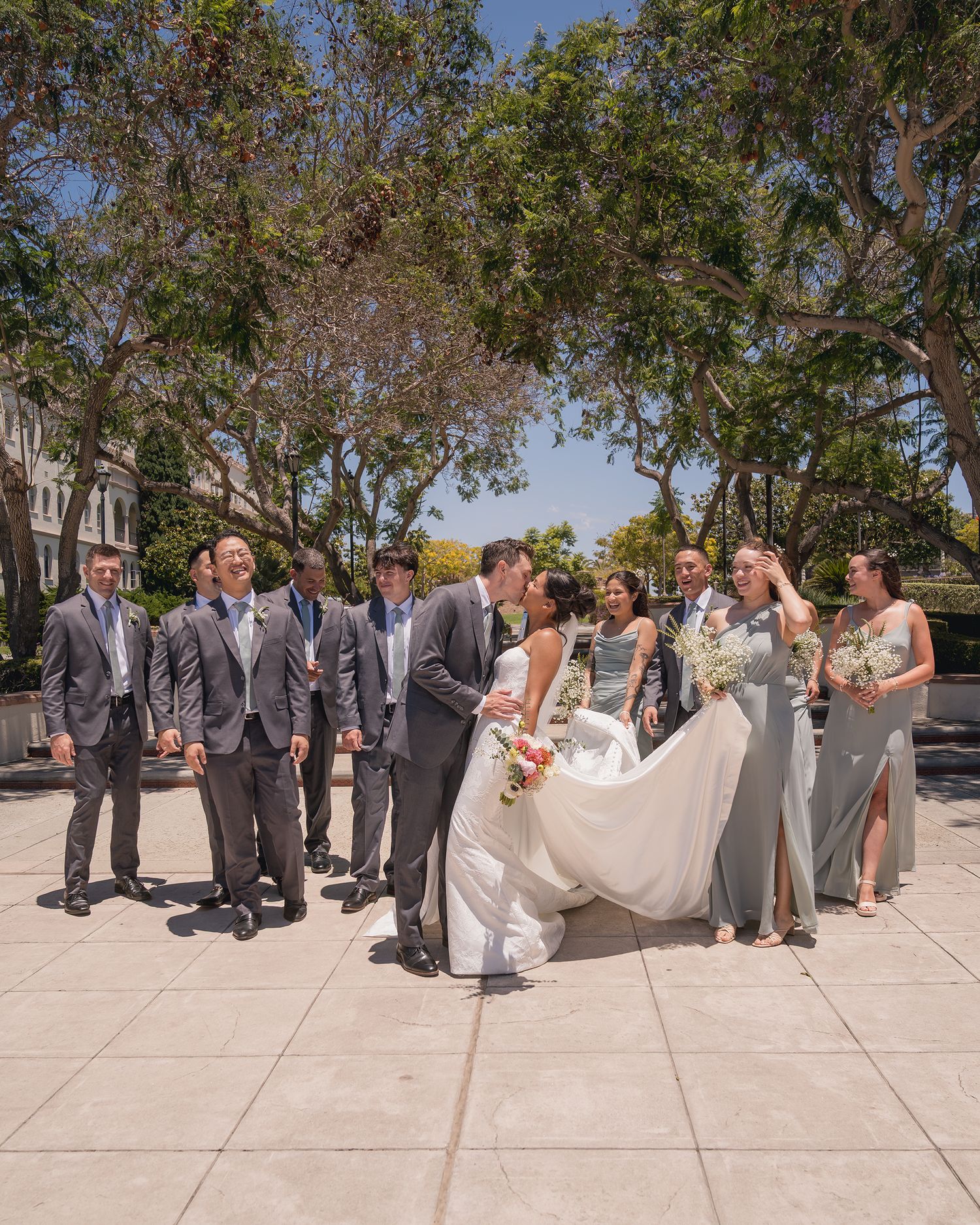 Wedding party with bride and groom kissing surrounded by groomsmen and bridesmaids in gray outdoors.