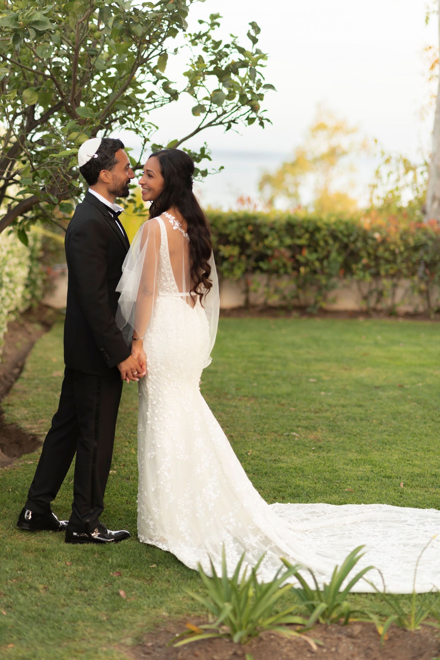 Bride in a lace mermaid gown with a long train holds hands with her groom in a black suit and kippah in a sunny garden.