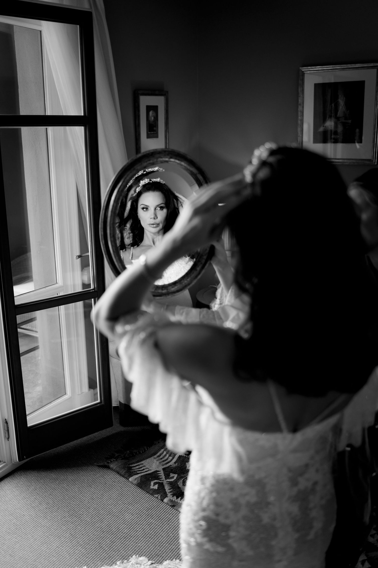 Black and white photo of a bride in a lace gown adjusting her hair while admiring her reflection in an oval mirror.