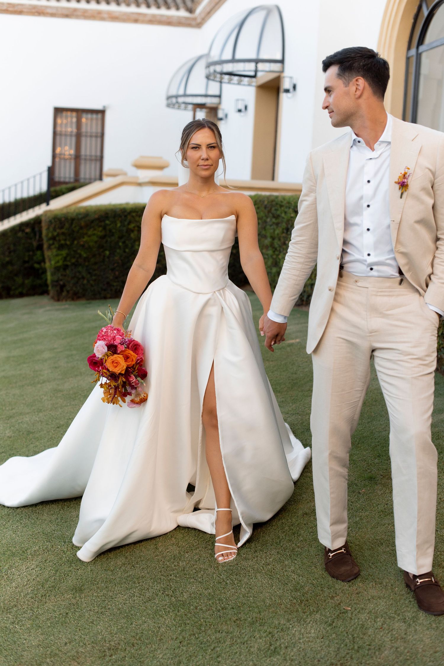 Bride in a strapless satin gown with a high slit holds a colorful bouquet and hands with a groom in a beige suit on a sunny lawn.