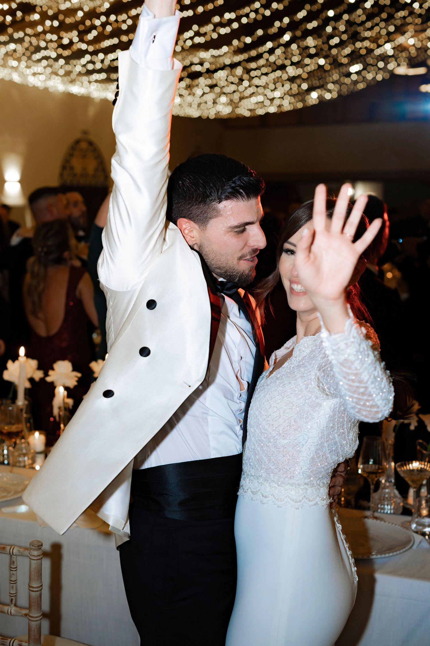 Bride and groom dance with raised hands at an elegant wedding reception under twinkling string lights and candlelit tables.