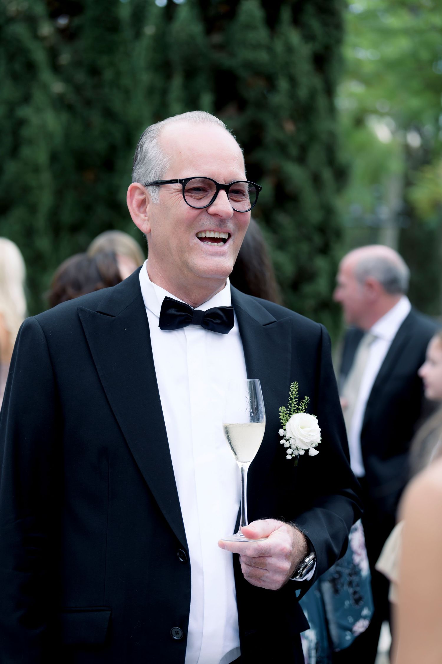 Smiling man in glasses, black tuxedo and bow tie holds a champagne flute at an outdoor wedding reception with guests behind him.