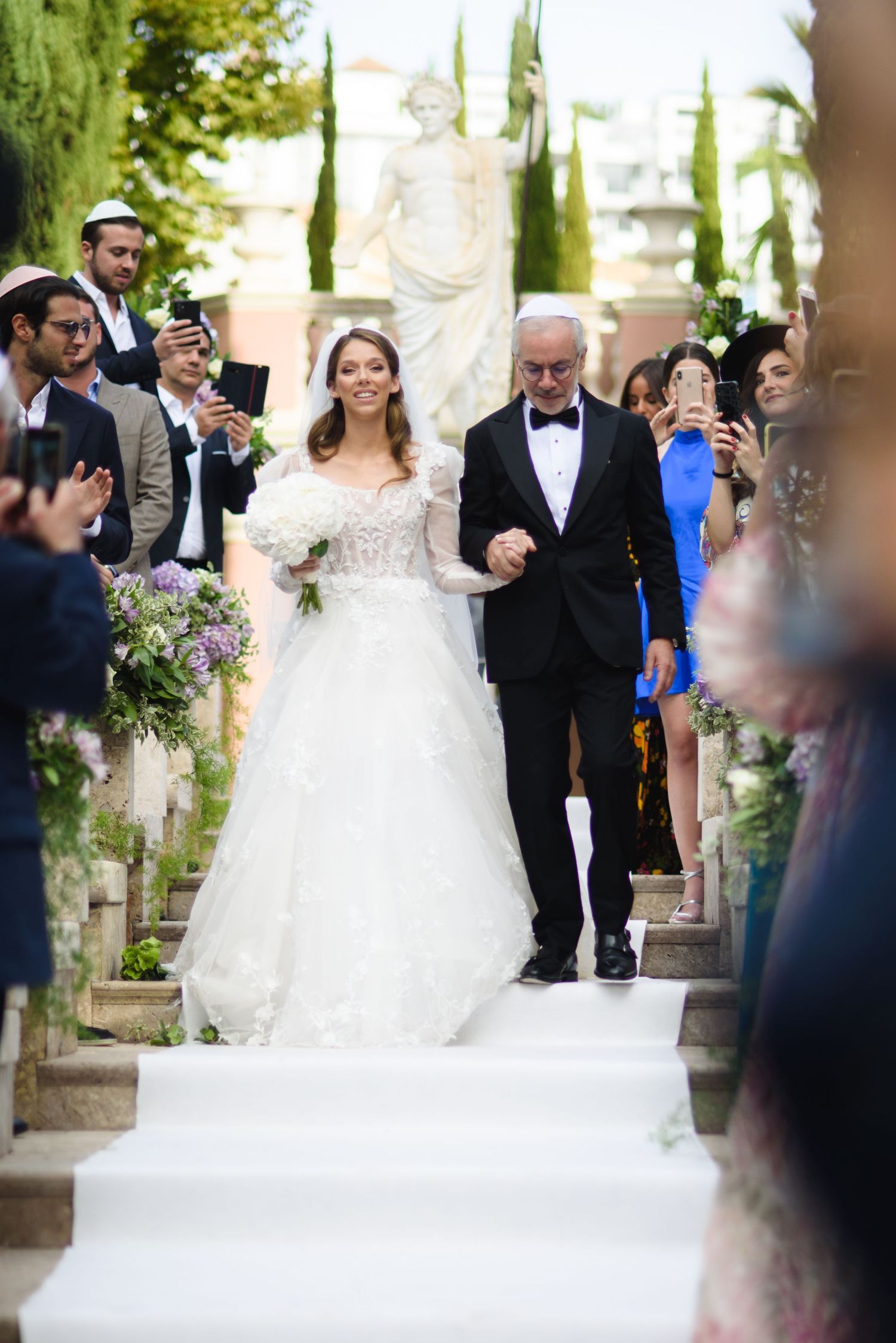 Bride in a lace ball gown holding a bouquet walks down white-carpeted steps arm in arm with a man in a tuxedo as guests watch.