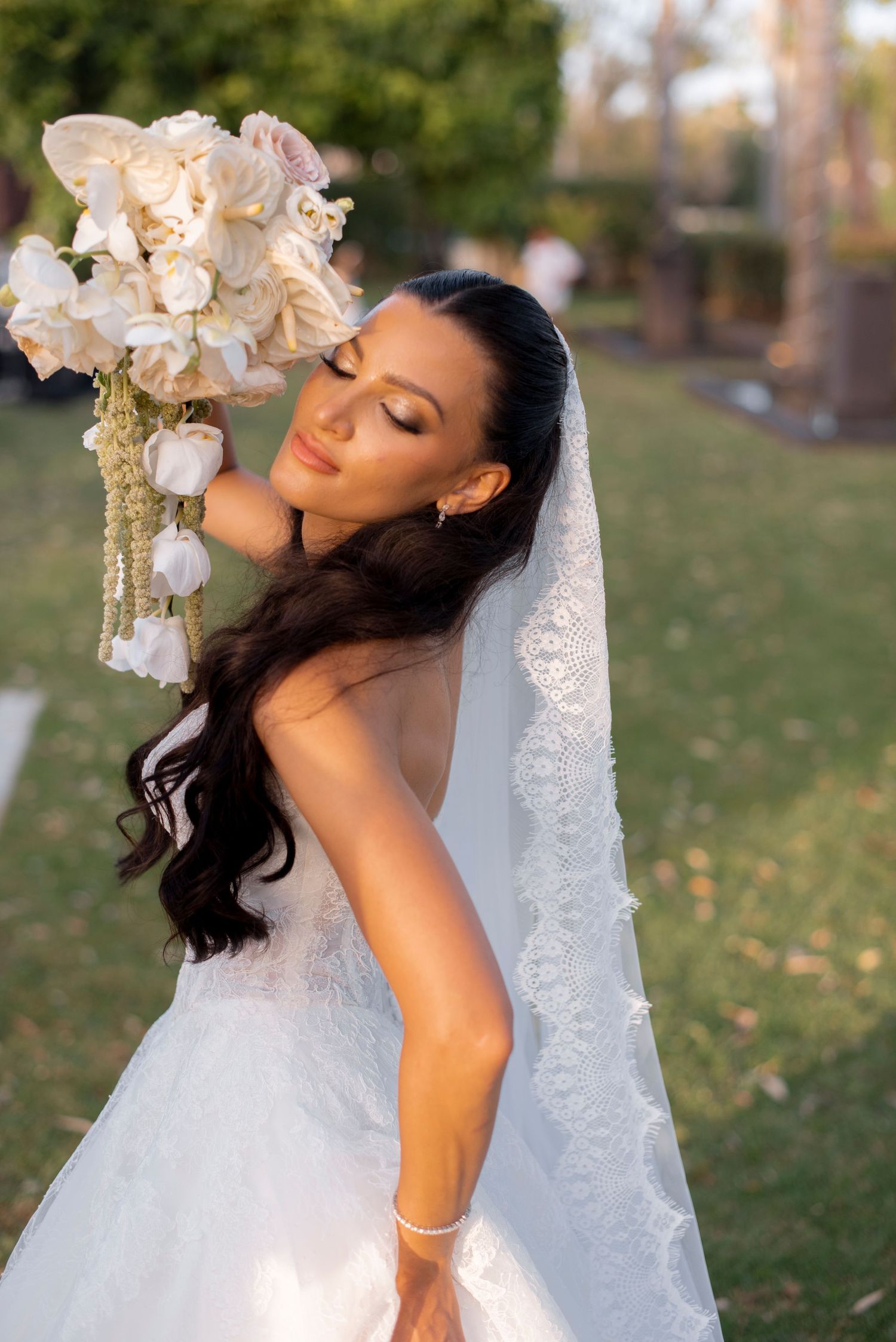Bride in a strapless white lace gown and long veil holds a cascading white orchid bouquet while posing in a sunlit garden.