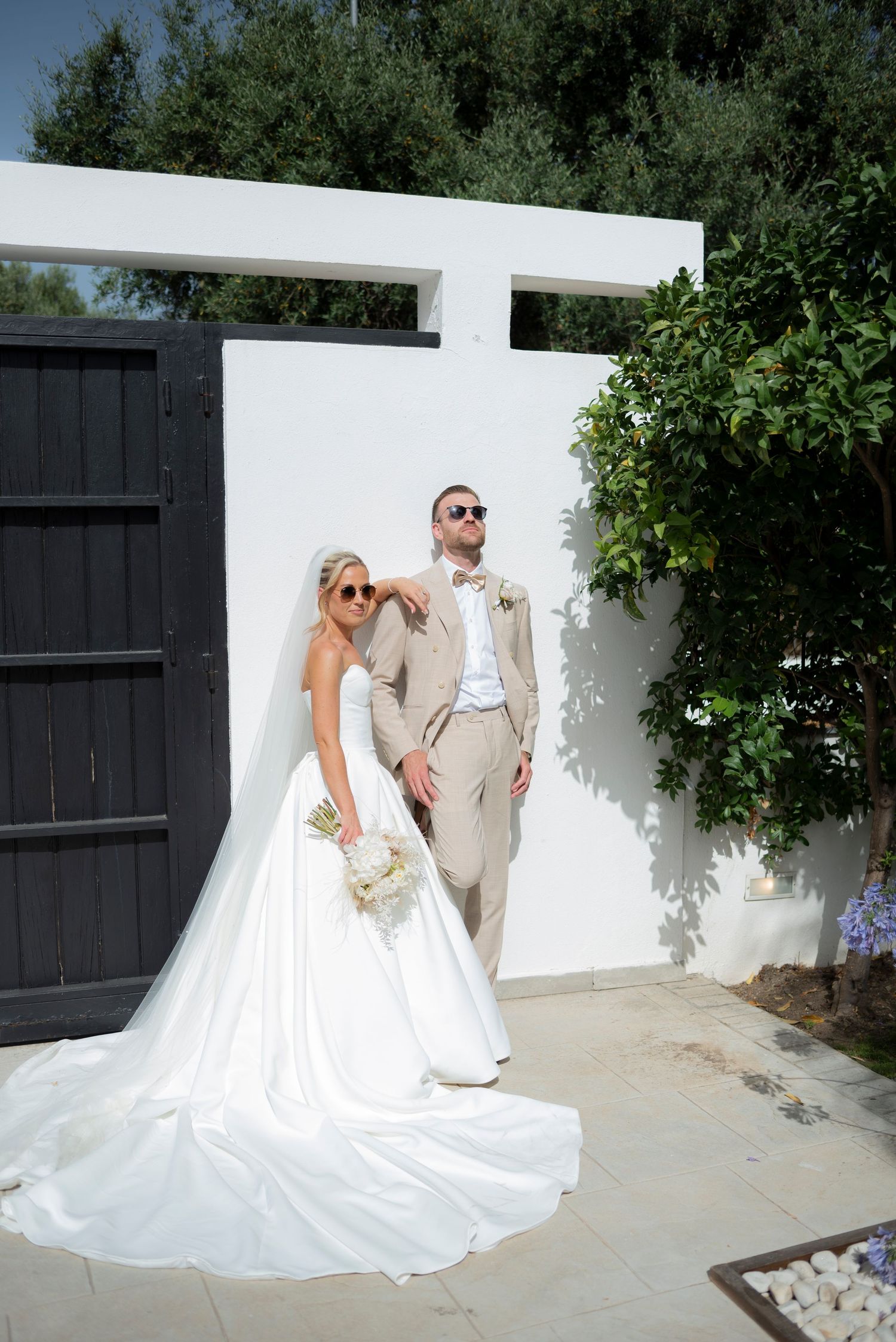 Bride in a flowing white gown and veil poses with a groom in a tan suit and sunglasses against a white wall near olive trees.