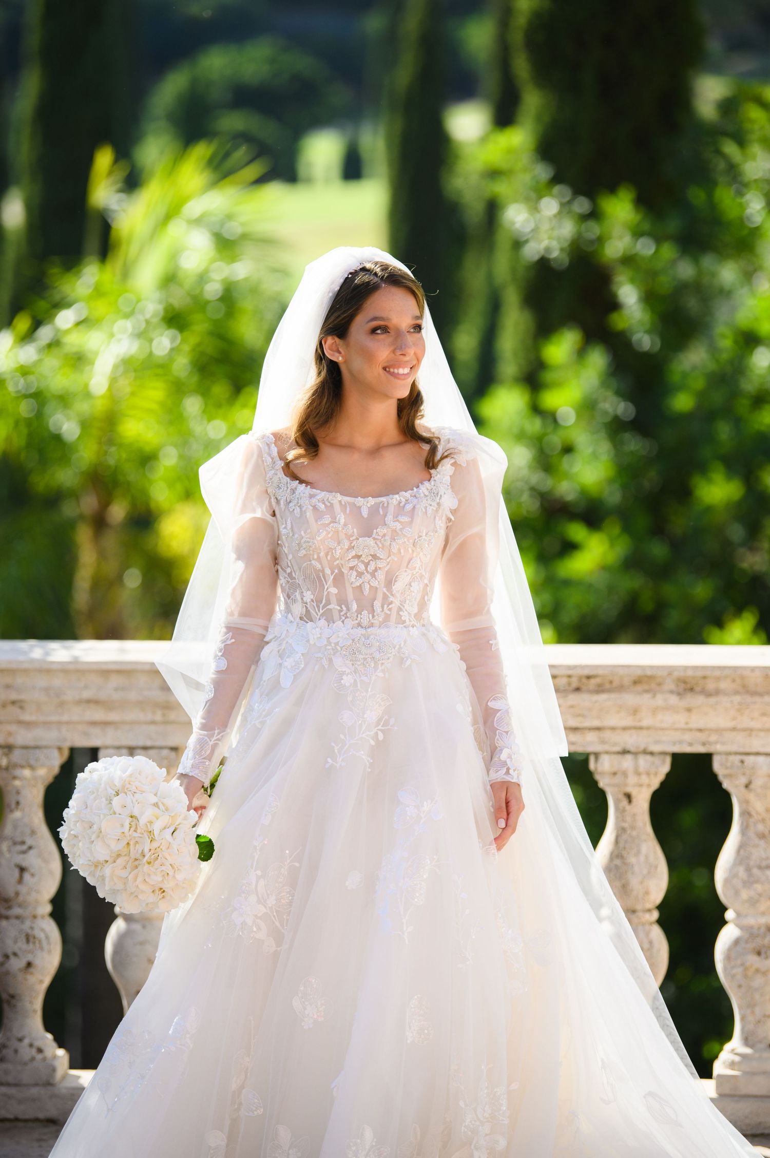 Smiling bride in a lace long-sleeve ball gown and veil holding a white bouquet on a stone balcony.