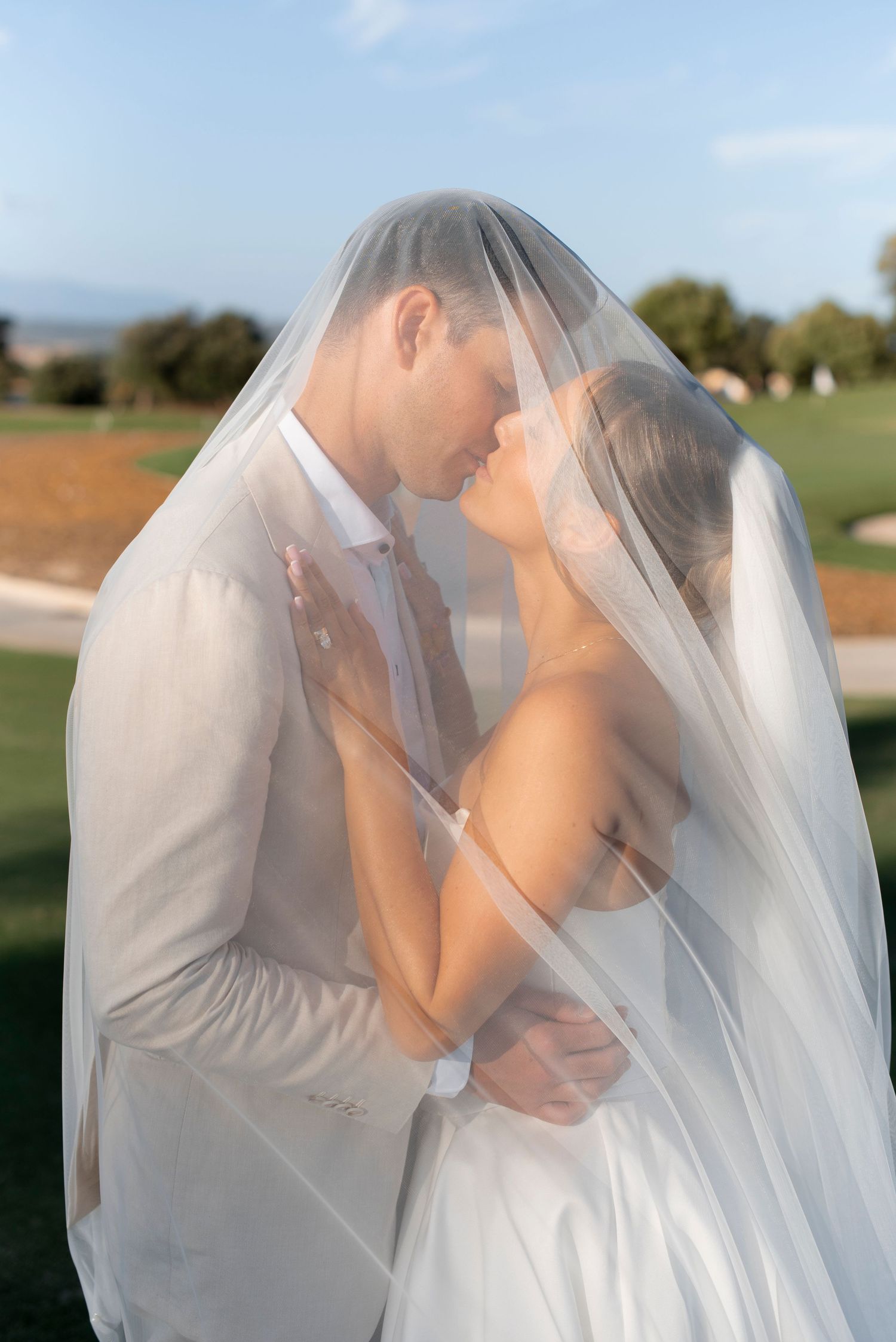 Bride and groom share a romantic kiss beneath a sheer wedding veil on a sunny golf course, with trees and blue sky behind them.