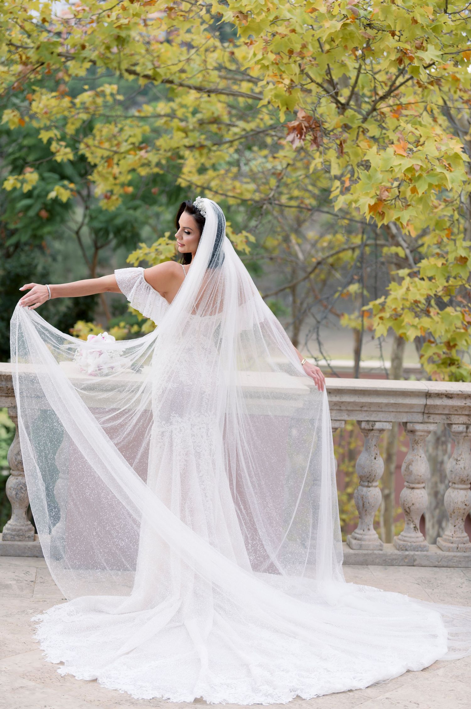 Bride in a lace mermaid wedding gown and long flowing veil poses on a stone terrace beside autumn trees.