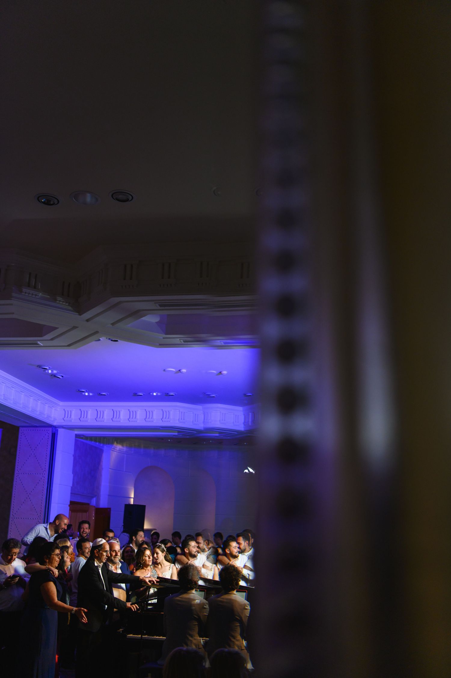 Crowd of guests gathered in an elegant ballroom with ornate ceiling details and purple lighting, seen through a dark foreground frame.