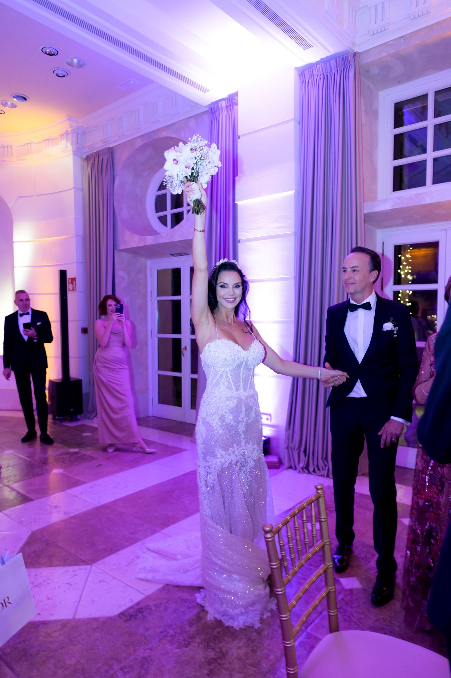Bride in a strapless lace gown raises her bouquet high while holding hands with a groom in a black tuxedo at a reception.