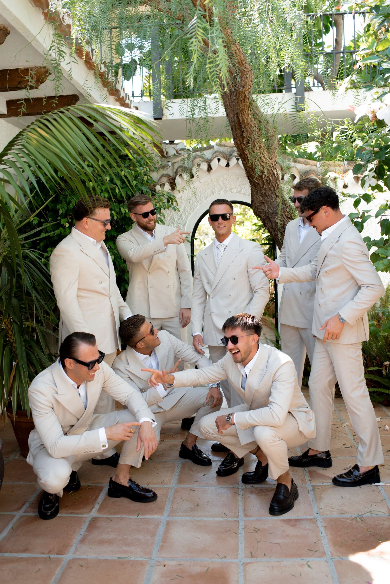 Groomsmen in cream suits and sunglasses pose playfully in a sunlit terracotta courtyard surrounded by greenery at a wedding.