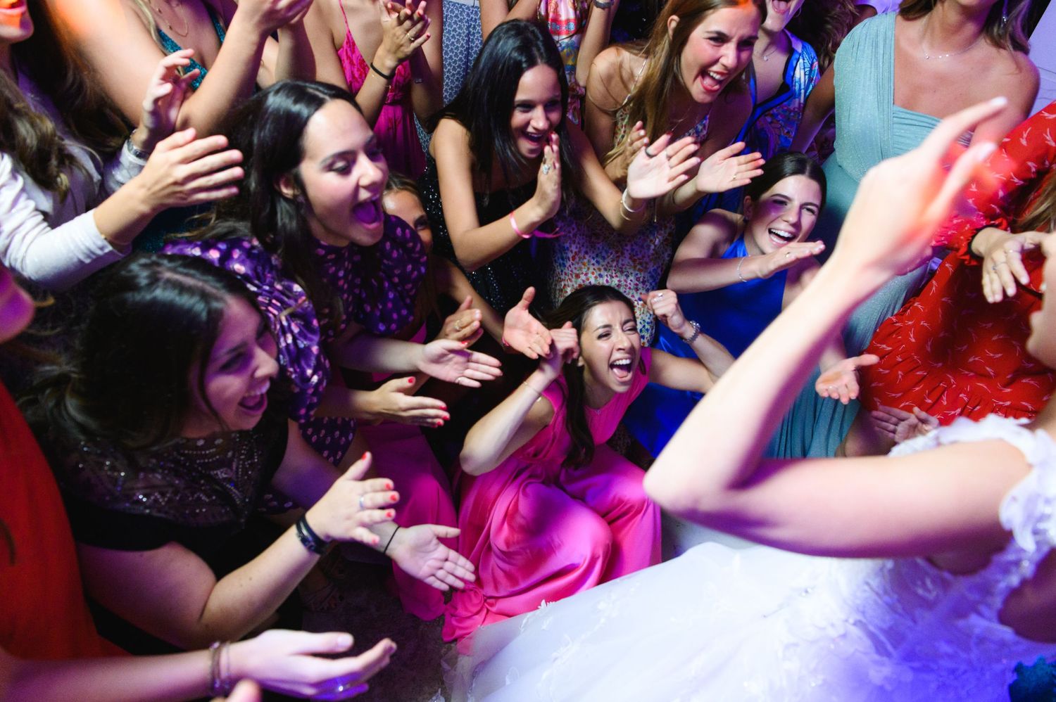 Guests clap and cheer in a lively circle around a dancing bride in a pink dress at a wedding reception.