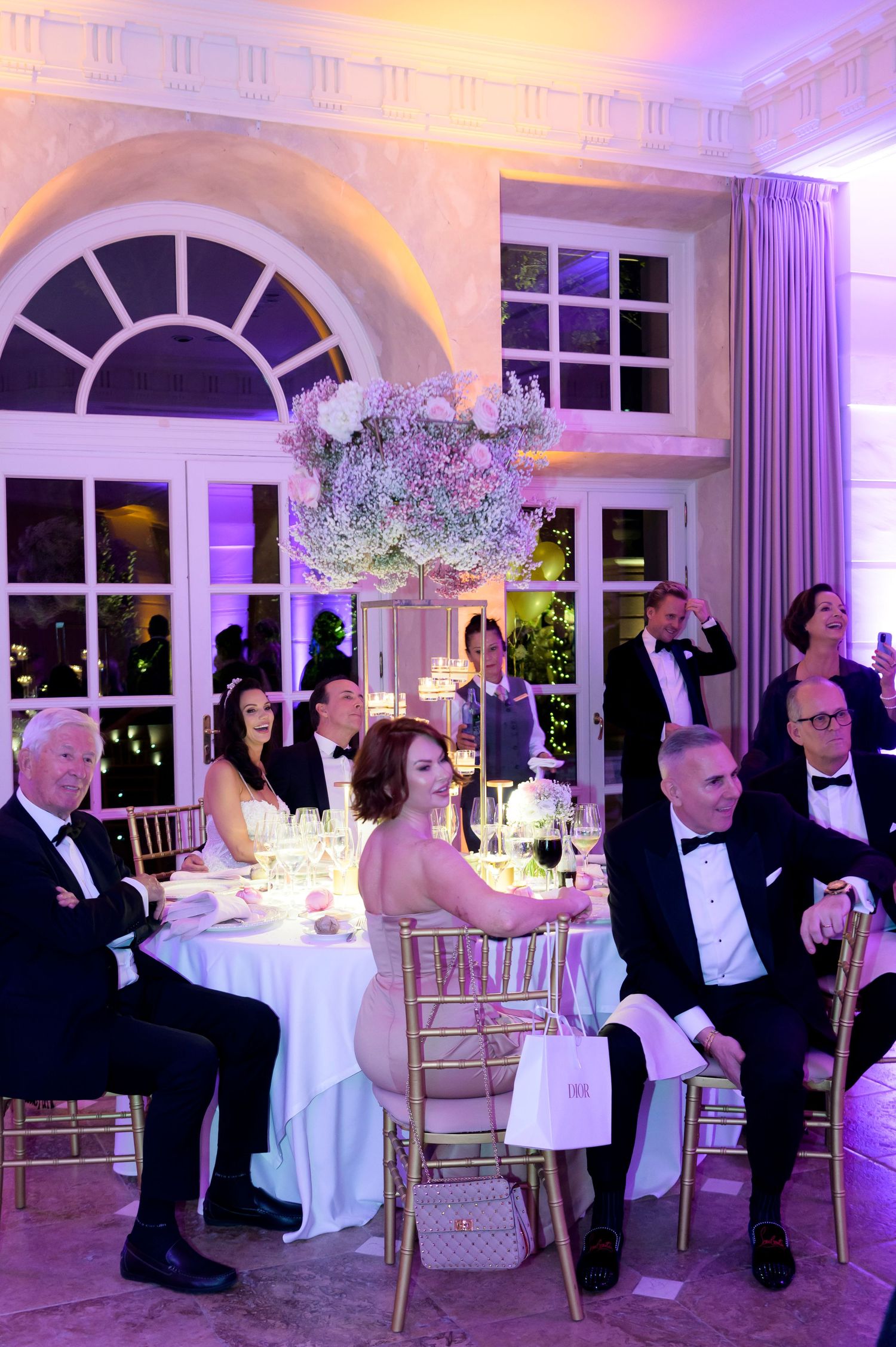 Formally dressed wedding guests sit at a white-clothed table beneath a tall floral centerpiece in a purple-lit ballroom.