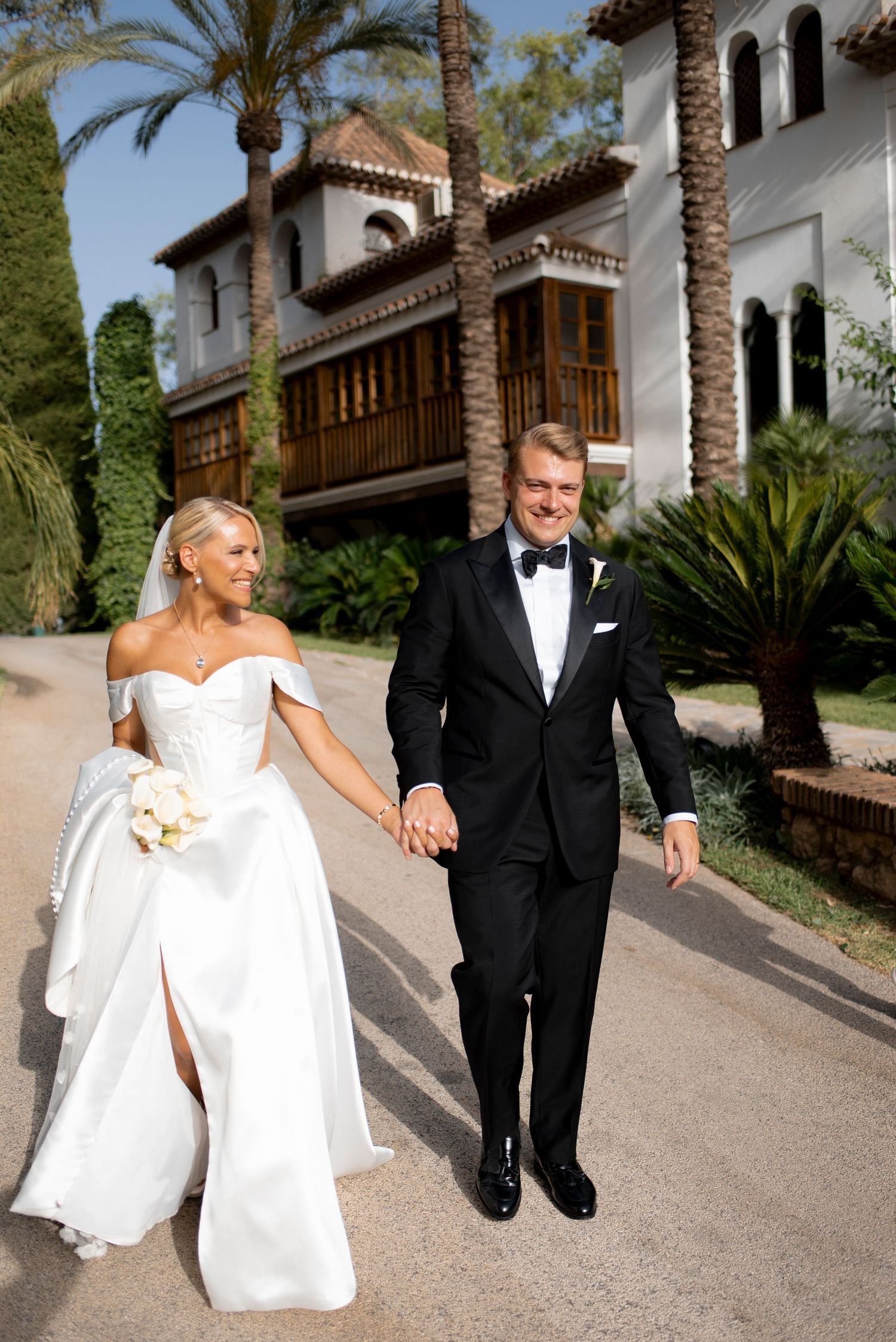 Bride in a white off-the-shoulder gown and groom in a black tuxedo hold hands walking past palm trees at a Mediterranean-style venue.
