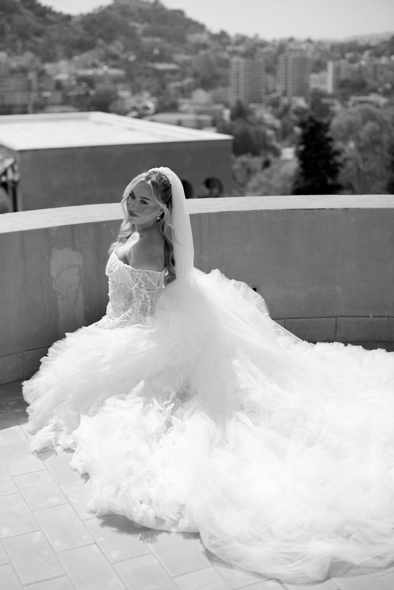 Black and white photo of a bride in a lace off-shoulder gown with a voluminous tulle train seated on a rooftop terrace overlooking the city.
