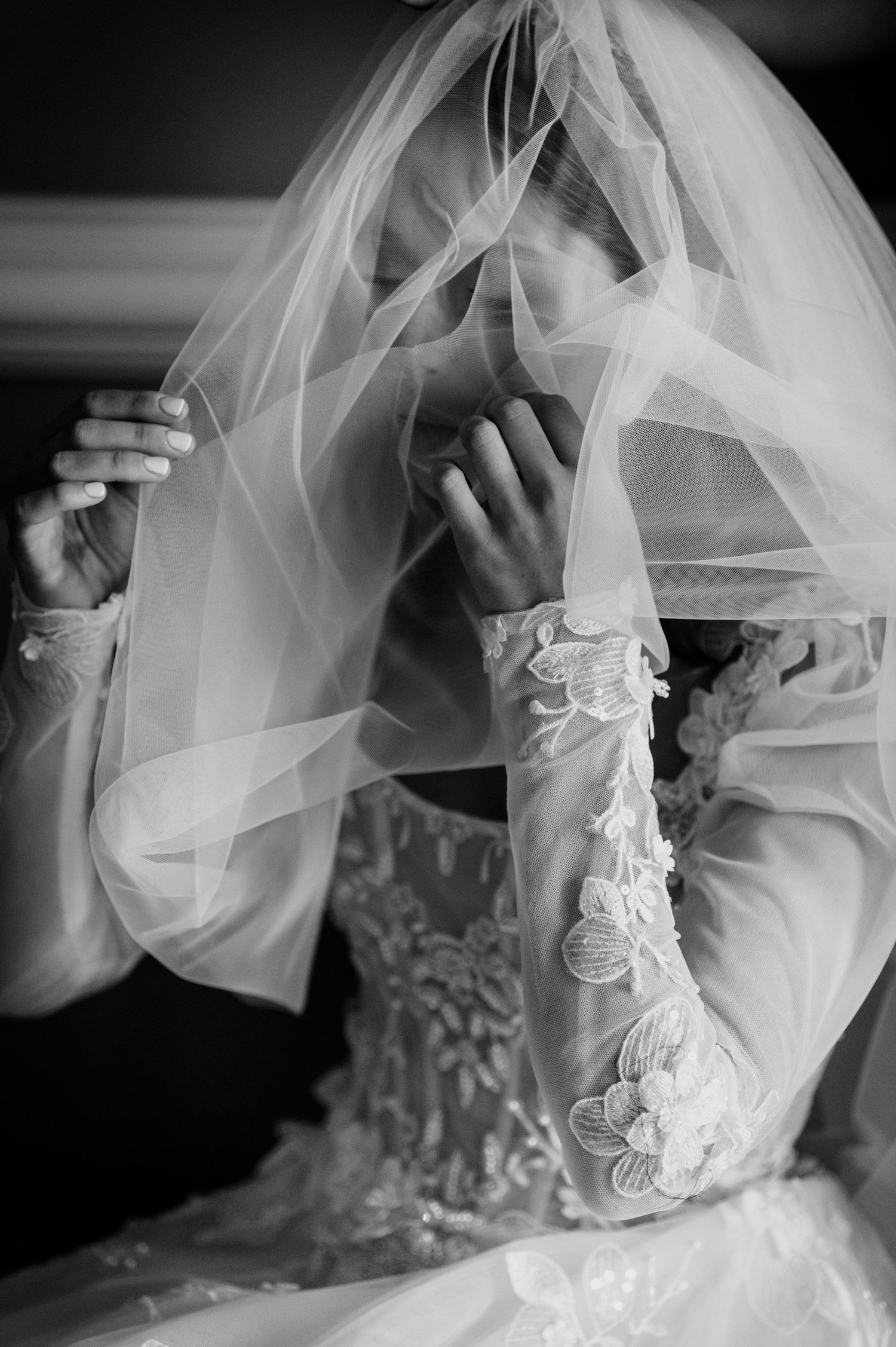 Black and white photo of a bride in a lace wedding gown with a sheer veil covering her face as she raises her hands.