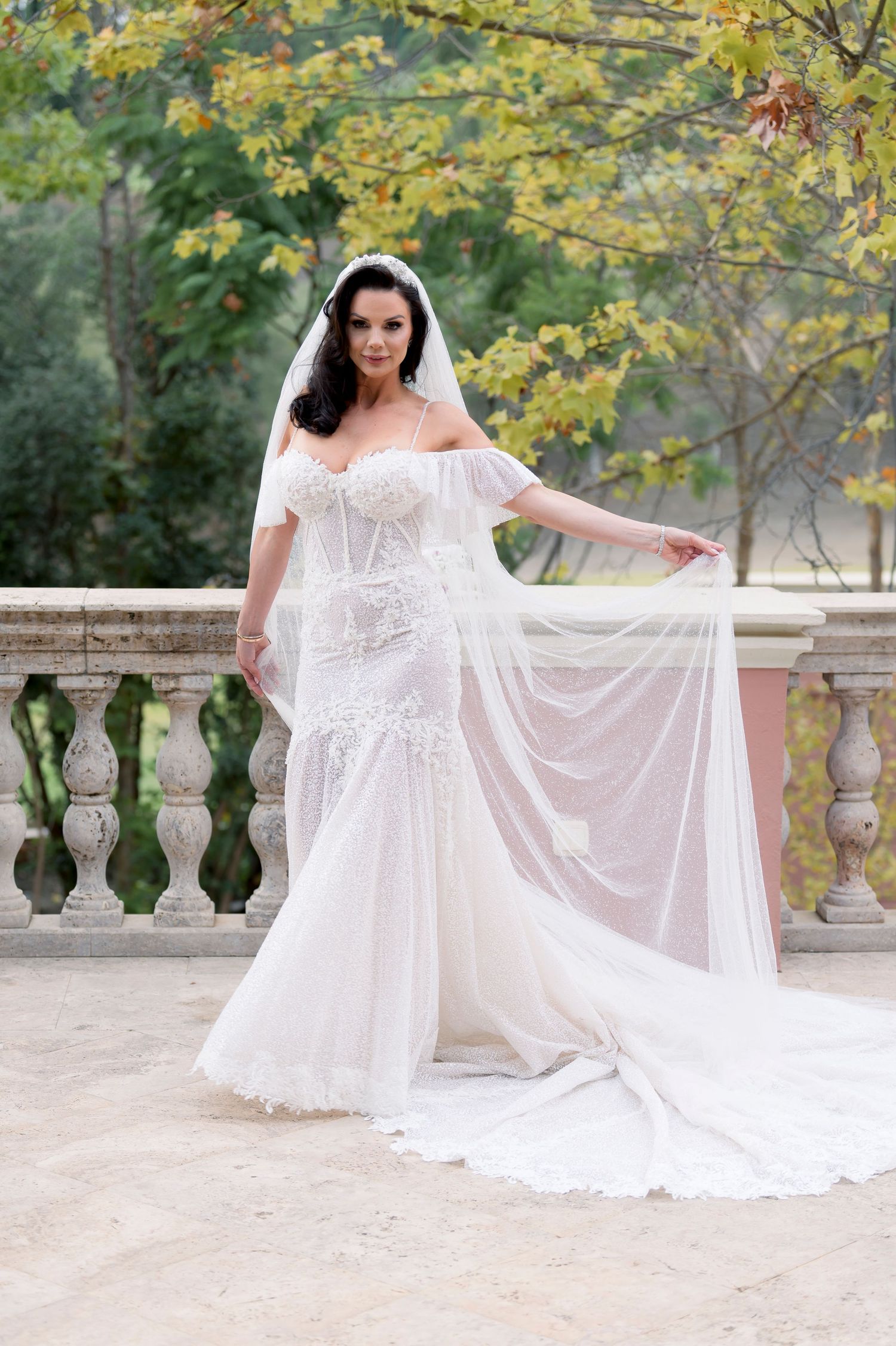 Bride in an off-the-shoulder lace mermaid wedding gown with a long flowing veil and train poses on a stone terrace beneath autumn trees.