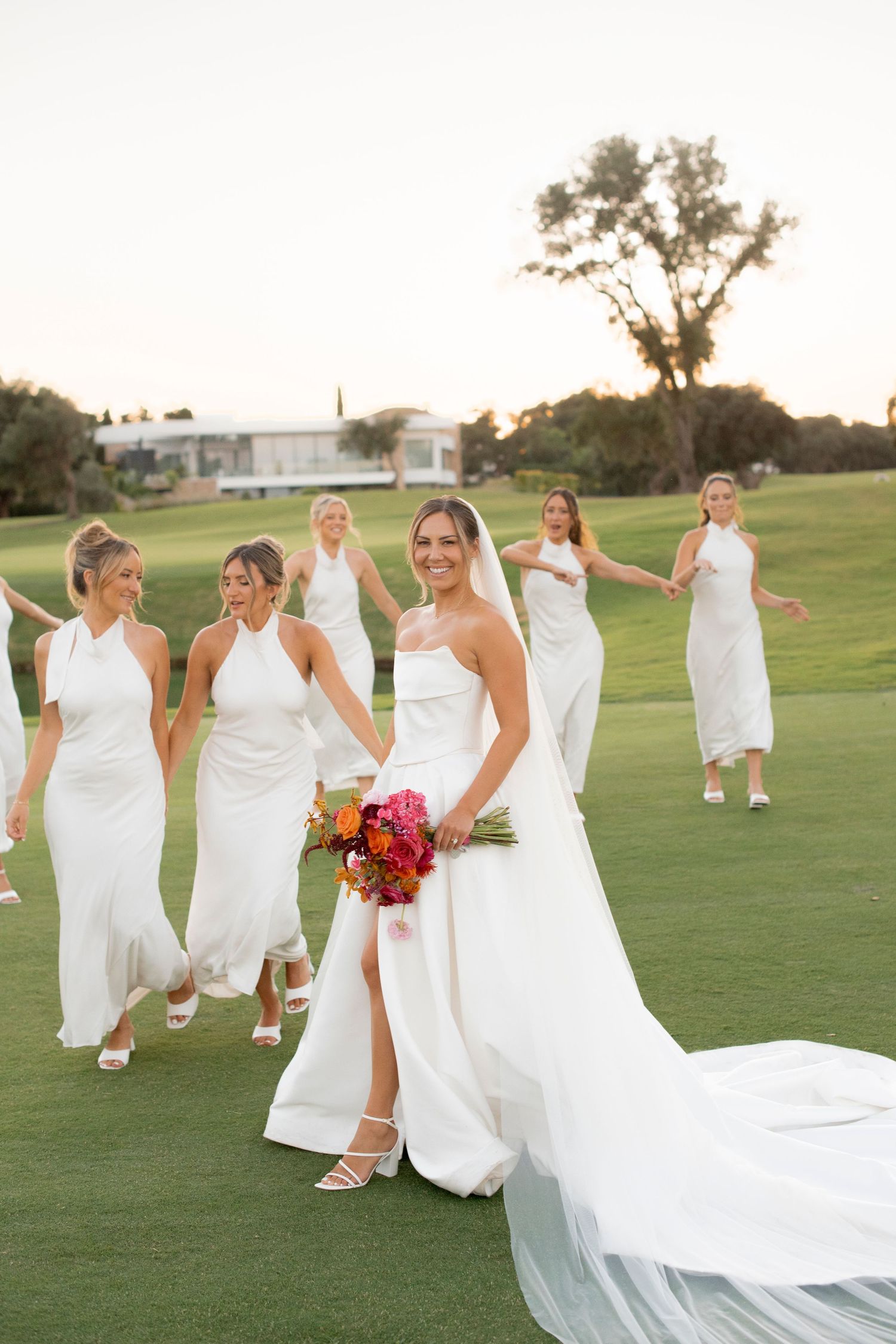 Bride in a white gown holding a colorful bouquet walks on a golf course lawn with bridesmaids in white dresses behind her at sunset.