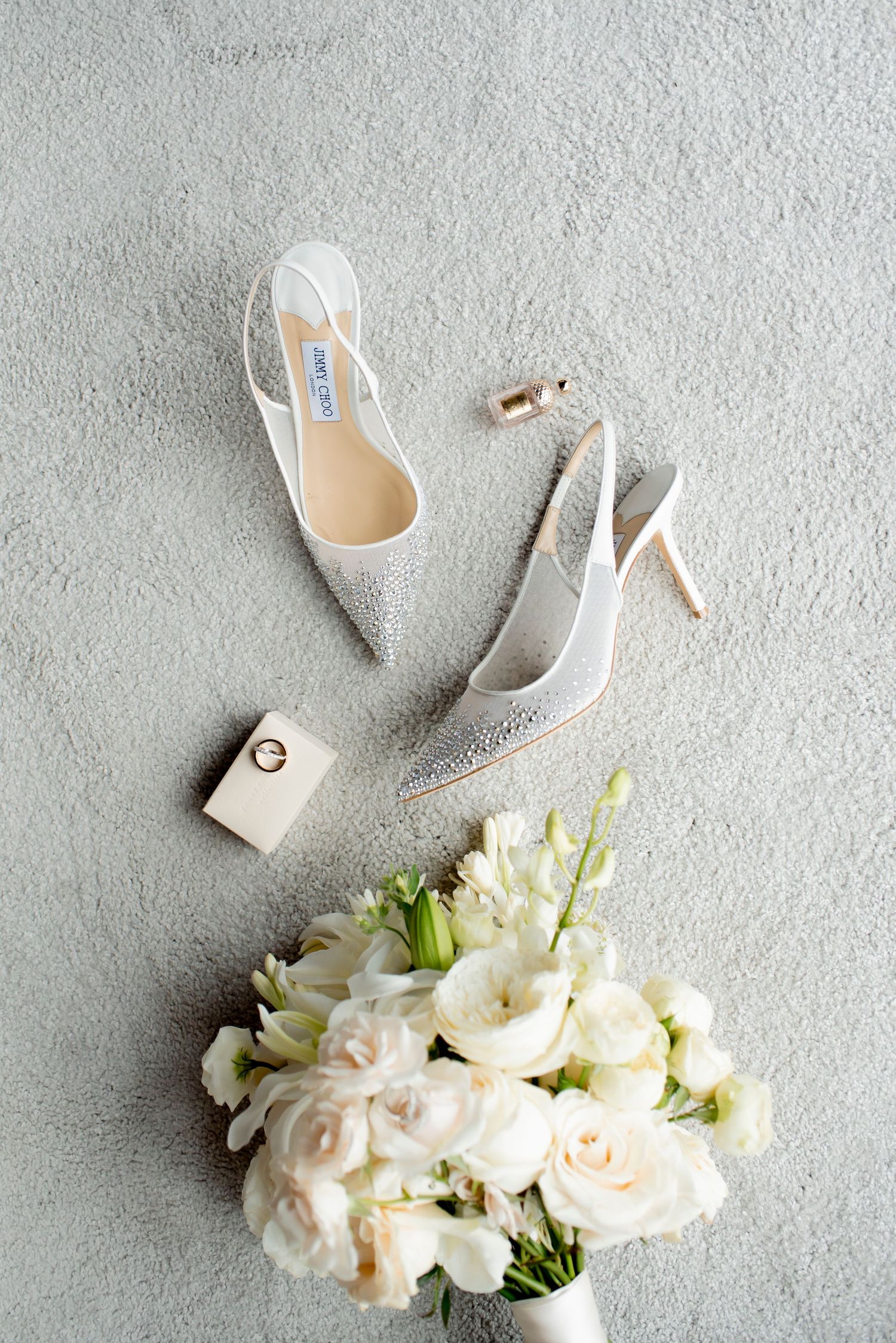 Sparkling white Jimmy Choo bridal heels, a ring box, a perfume vial and a white rose bouquet arranged on a gray textured surface.
