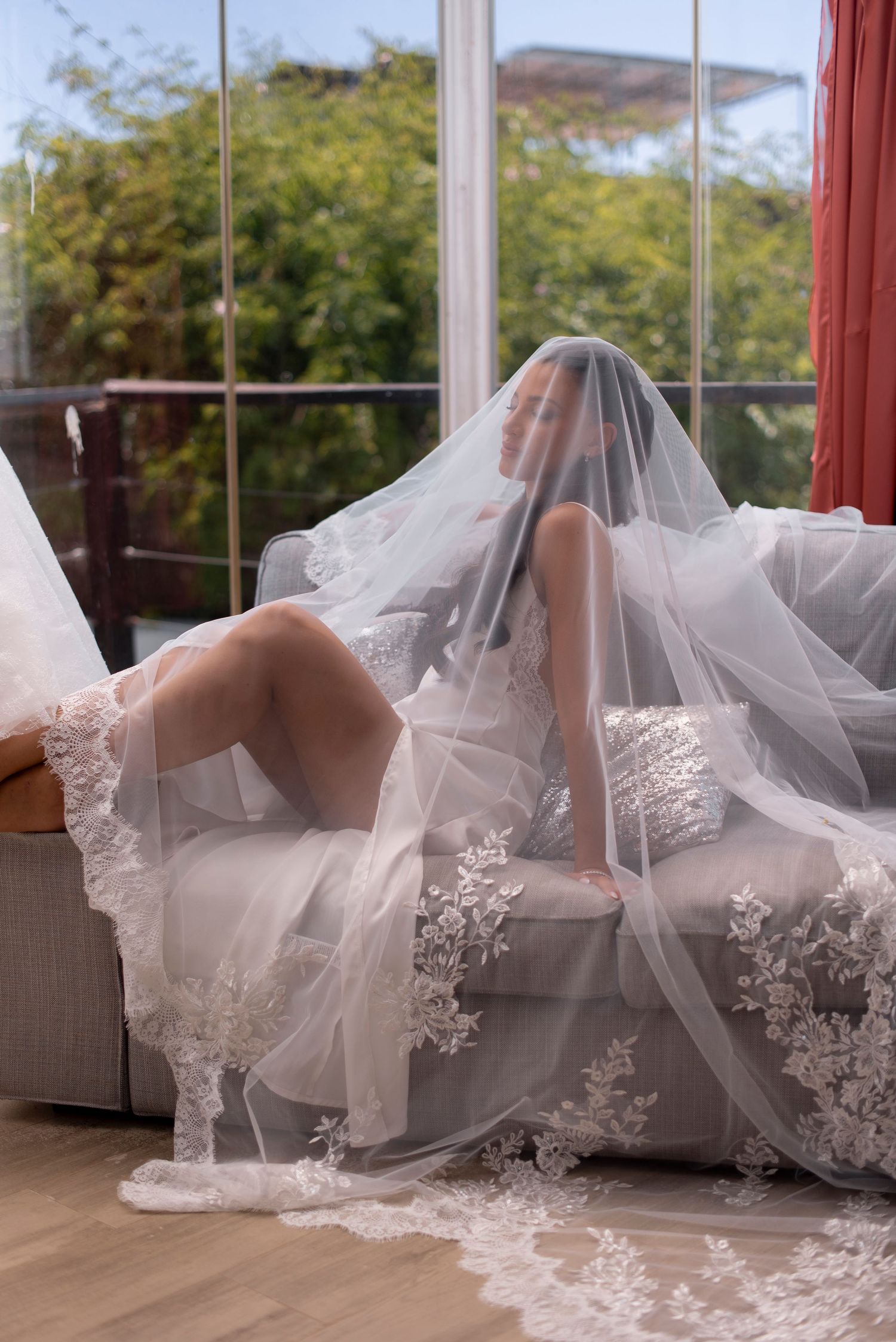 Bride reclining on a gray sofa beneath a sheer lace-trimmed bridal veil, with large windows and green trees behind her.