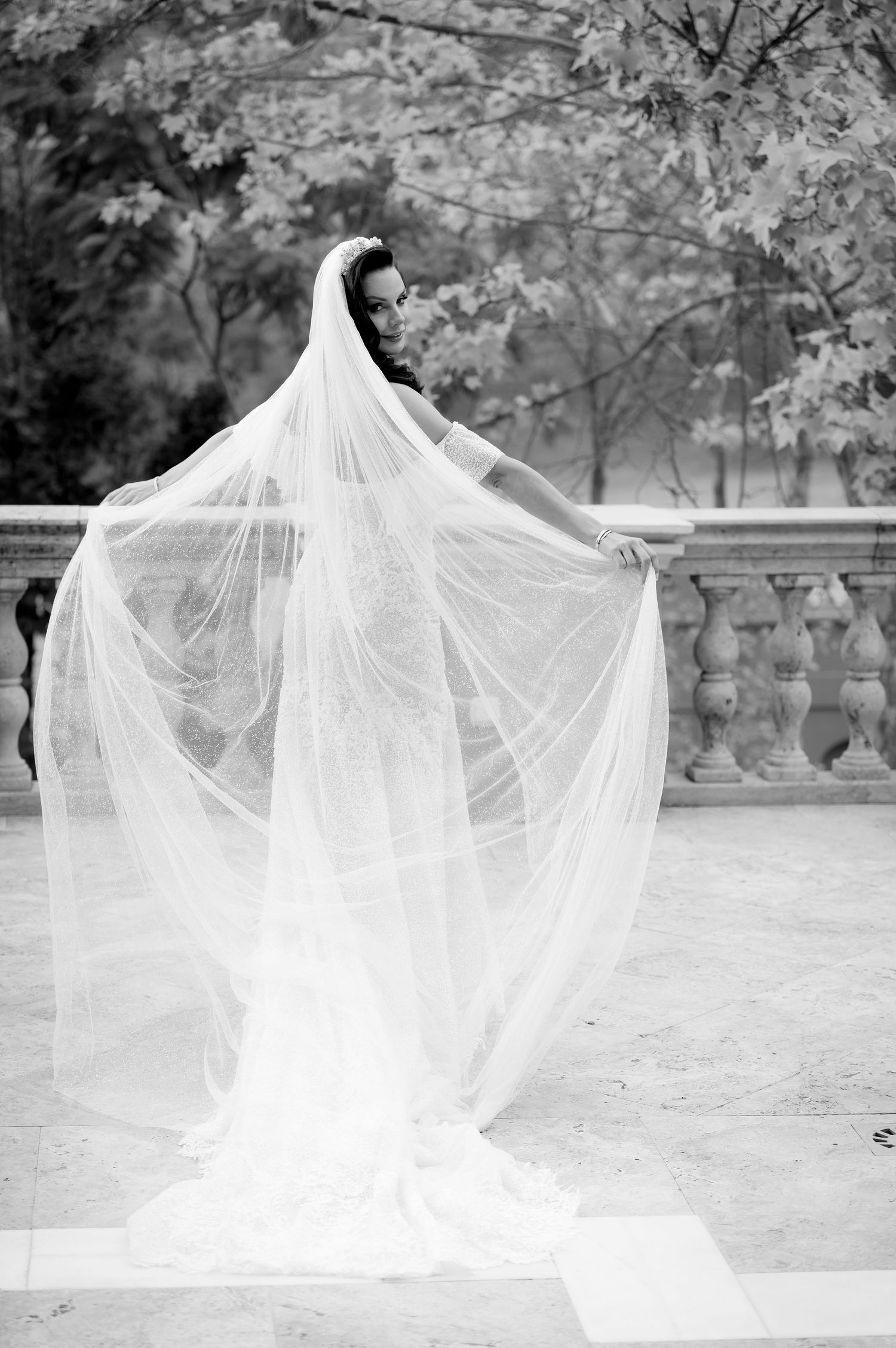 Black and white photo of a bride in a lace gown holding out a long sheer veil on a stone terrace with a balustrade and trees.
