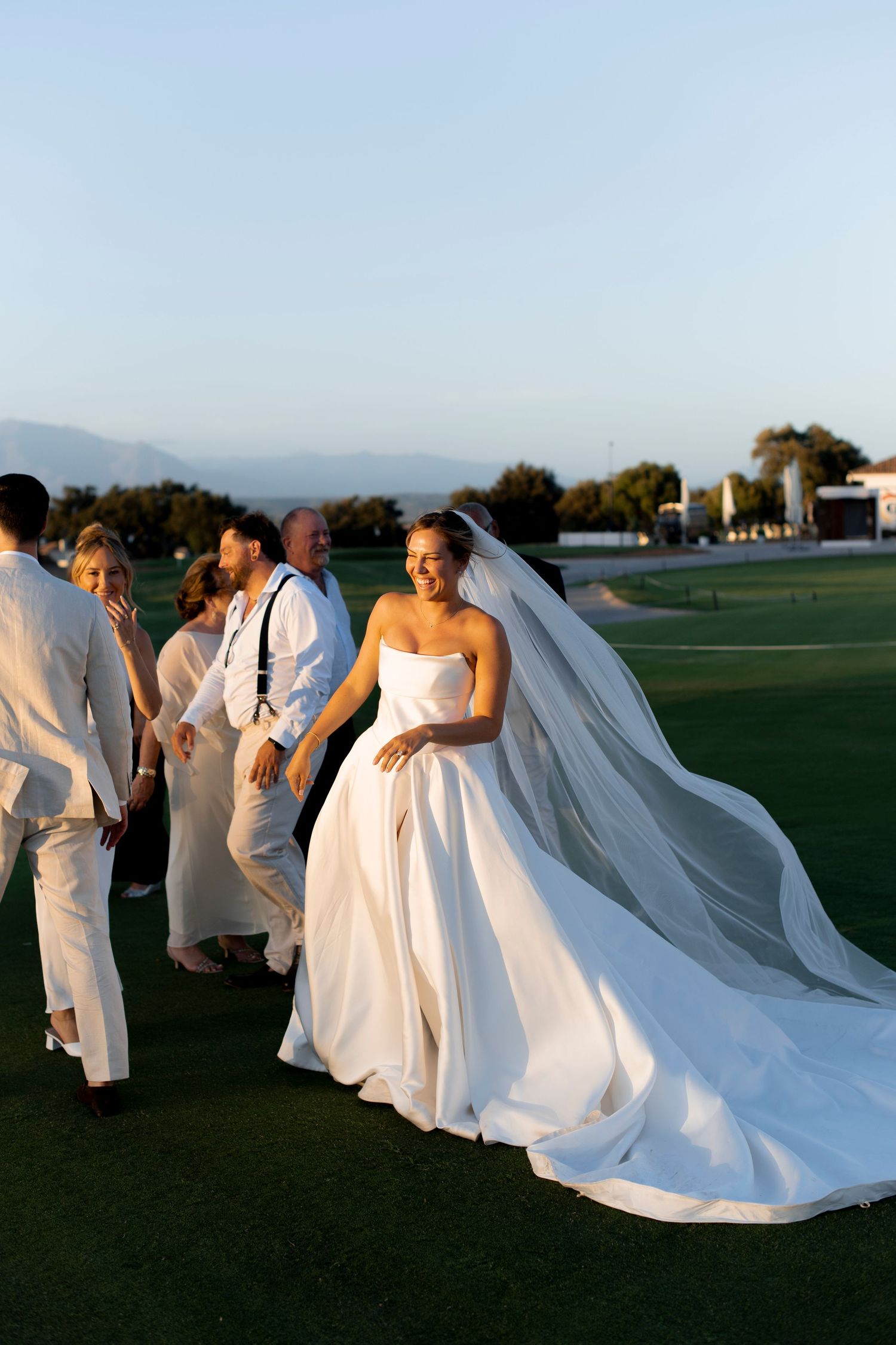 Bride in a strapless white satin gown and long veil walks across a golf course lawn at sunset with wedding guests behind her.